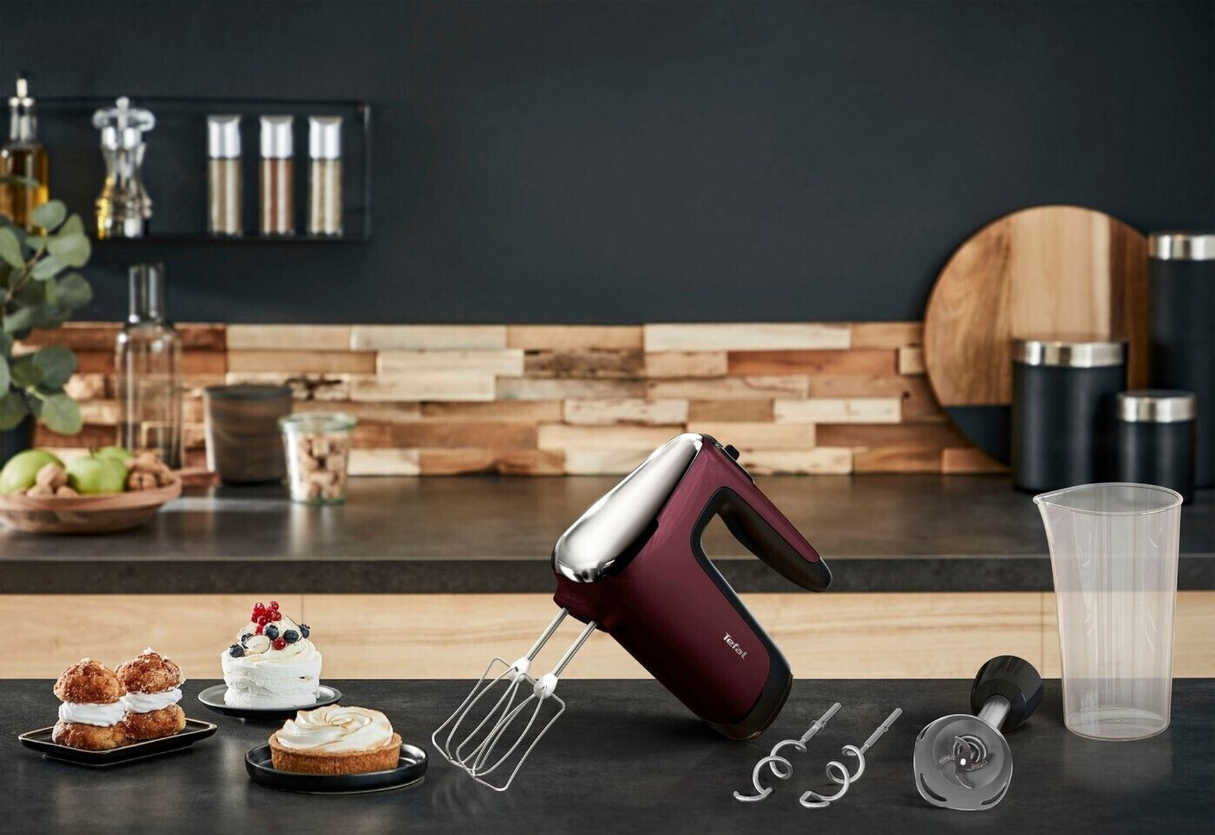 Una escena de cocina con una batidora de mano roja, ingredientes y postres sobre una encimera contra una pared oscura.