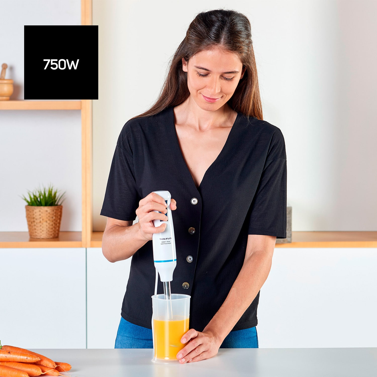 Kobieta miksuje sok pomarańczowy blenderem ręcznym w przezroczystym pojemniku. Kuchnia, biała powierzchnia.