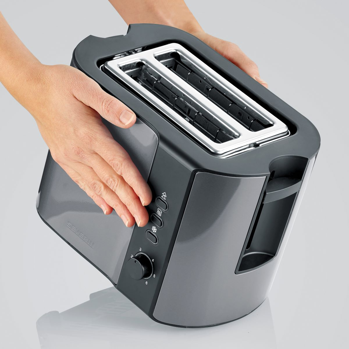 Dłonie trzymające szary toster, z przyciskami, na jasnoszarym tle.