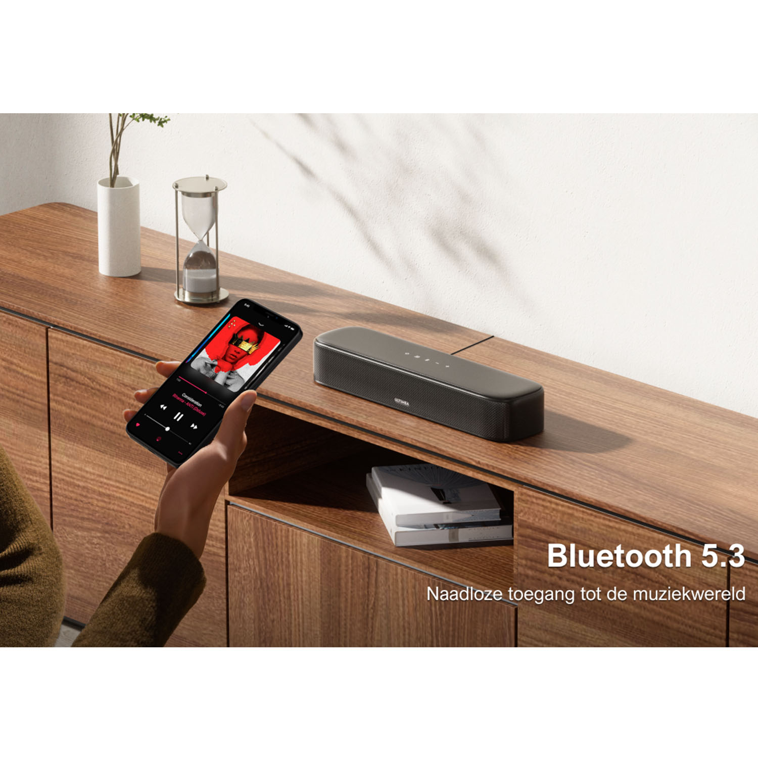 Hand holding a smartphone playing music, next to a soundbar on a wooden surface.
