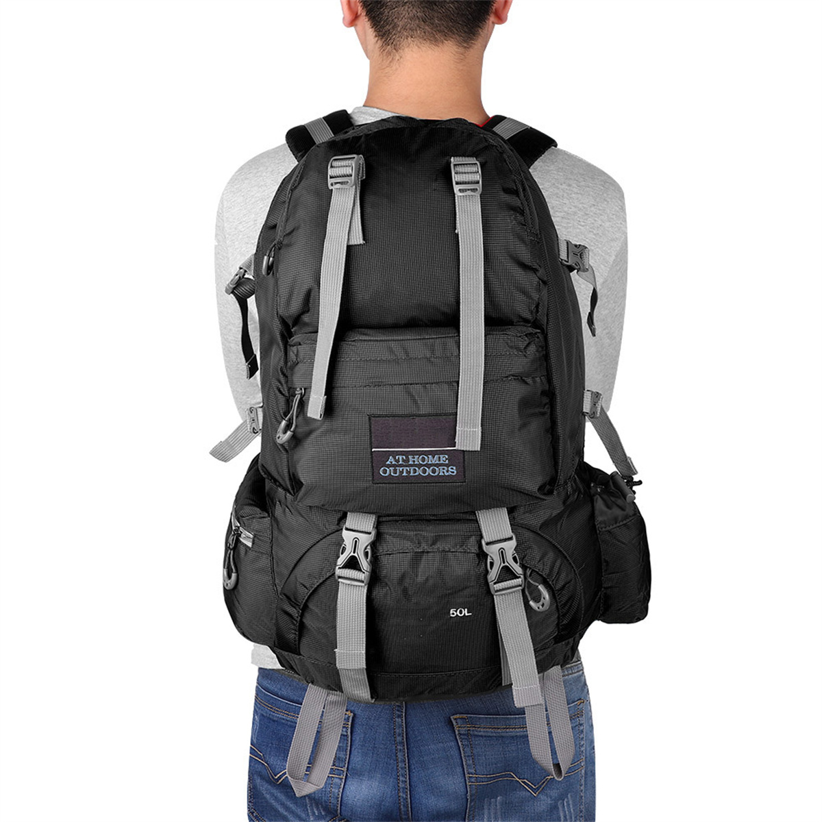 Person mit Rucksack. Schwarz mit grauen Riemen. Hat eine 50L-Markierung und einen 'AT HOME OUTDOORS'-Aufnäher.