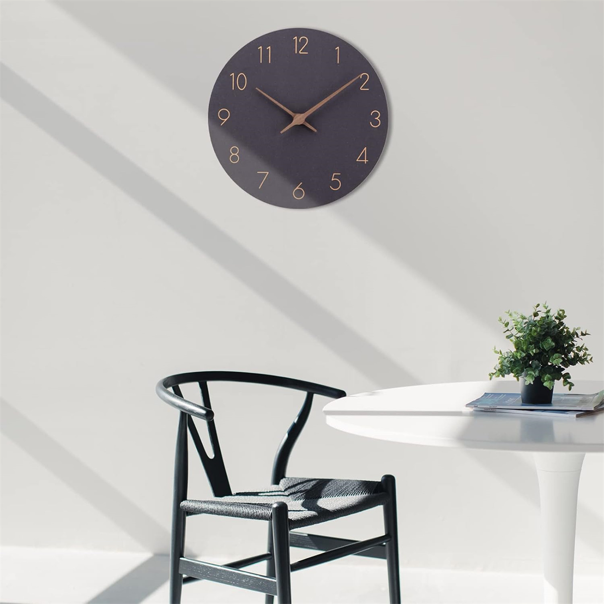 Eine dunkelgraue Uhr mit goldenen Zeigern und Ziffern hängt an einer weißen Wand; ein schwarzer Stuhl und Tisch im Vordergrund.