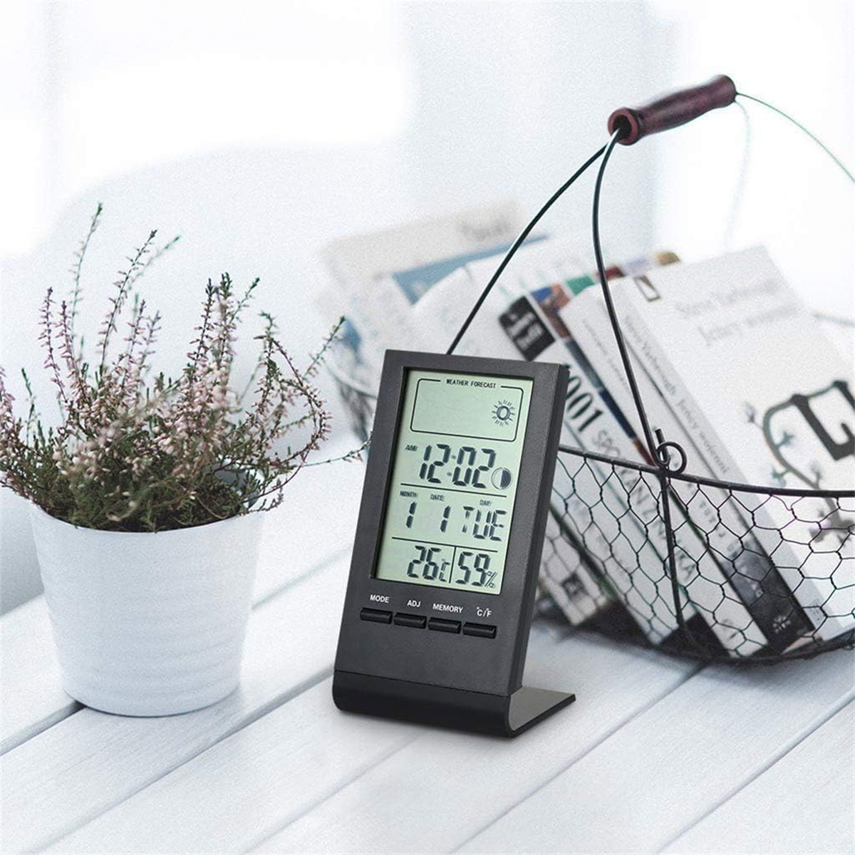 Schwarze Digitaluhr mit Blumentopf, Büchern in einem Korb und einem weißen Tisch.