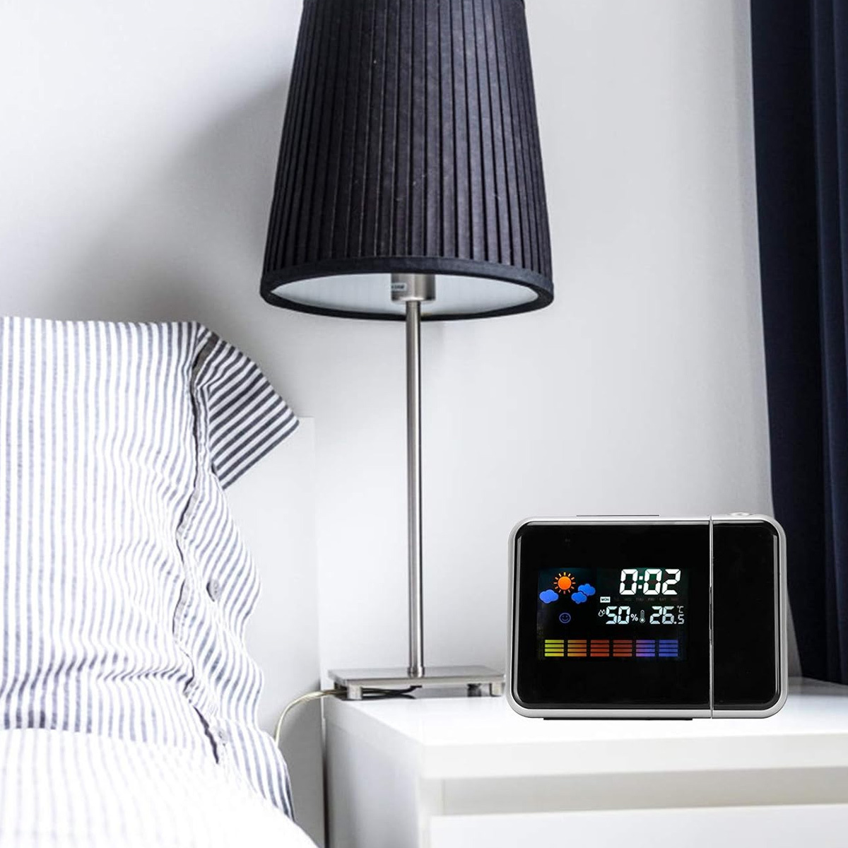 Eine Digitaluhr projiziert auf eine Wand. Sie zeigt Zeit, Wetter und Temperatur an. Im Hintergrund ist eine Lampe.
