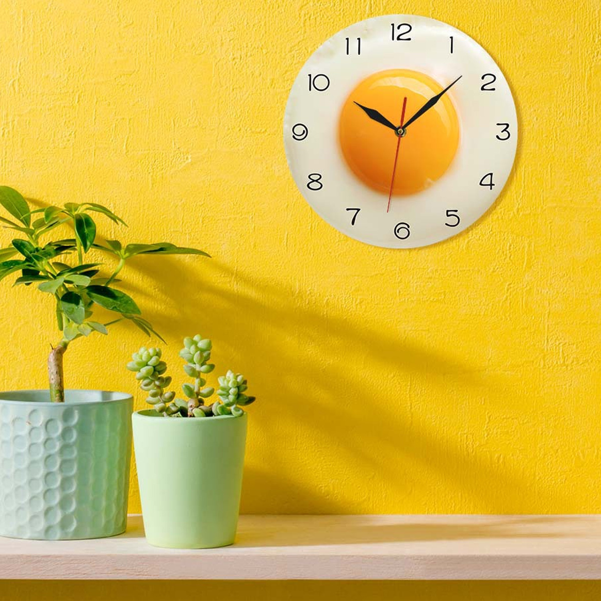 Eine Uhr mit Ei-Design an einer gelben Wand, mit Pflanzen auf einem Holzregal davor.