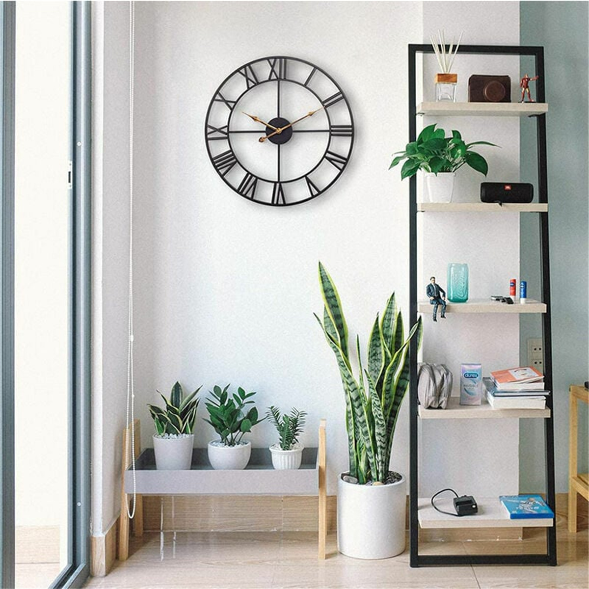 Eine schwarze Uhr mit römischen Ziffern hängt an einer weißen Wand über einem Regal mit Pflanzen und Dekoration.
