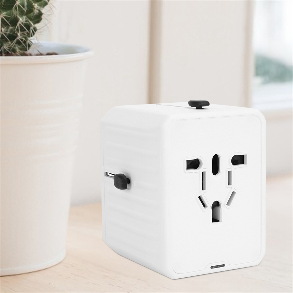 Ein weißer Universal-Reiseadapter mit mehreren Stecker-Konfigurationen. Im Hintergrund eine kleine Pflanze.