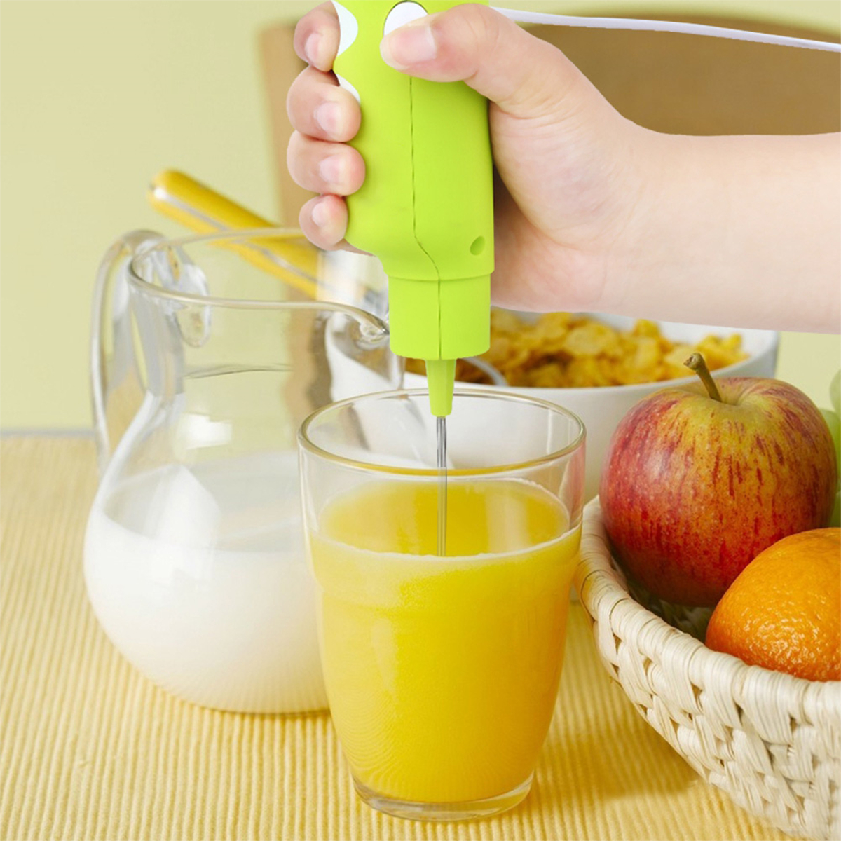 Hand hält einen grünen Aufschäumer, der gelbe Flüssigkeit in einem Glas mischt, mit Krug und Obst im Hintergrund.