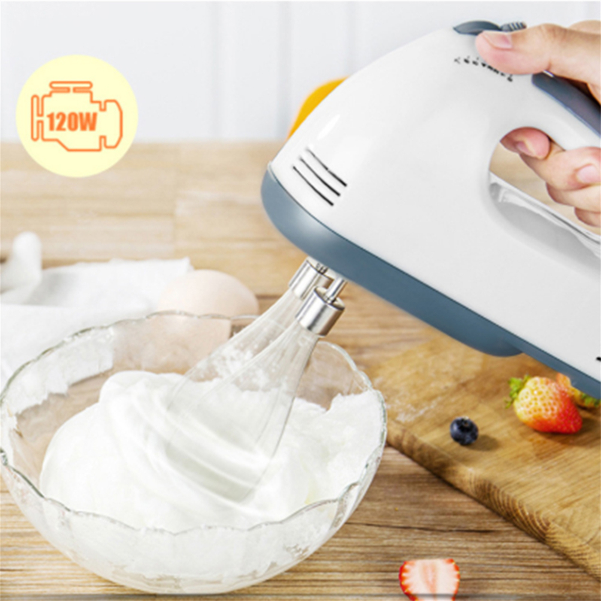 Ein weißer Handmixer verrührt Schlagsahne in einer Glasschüssel. Beeren und Erdbeeren liegen daneben.