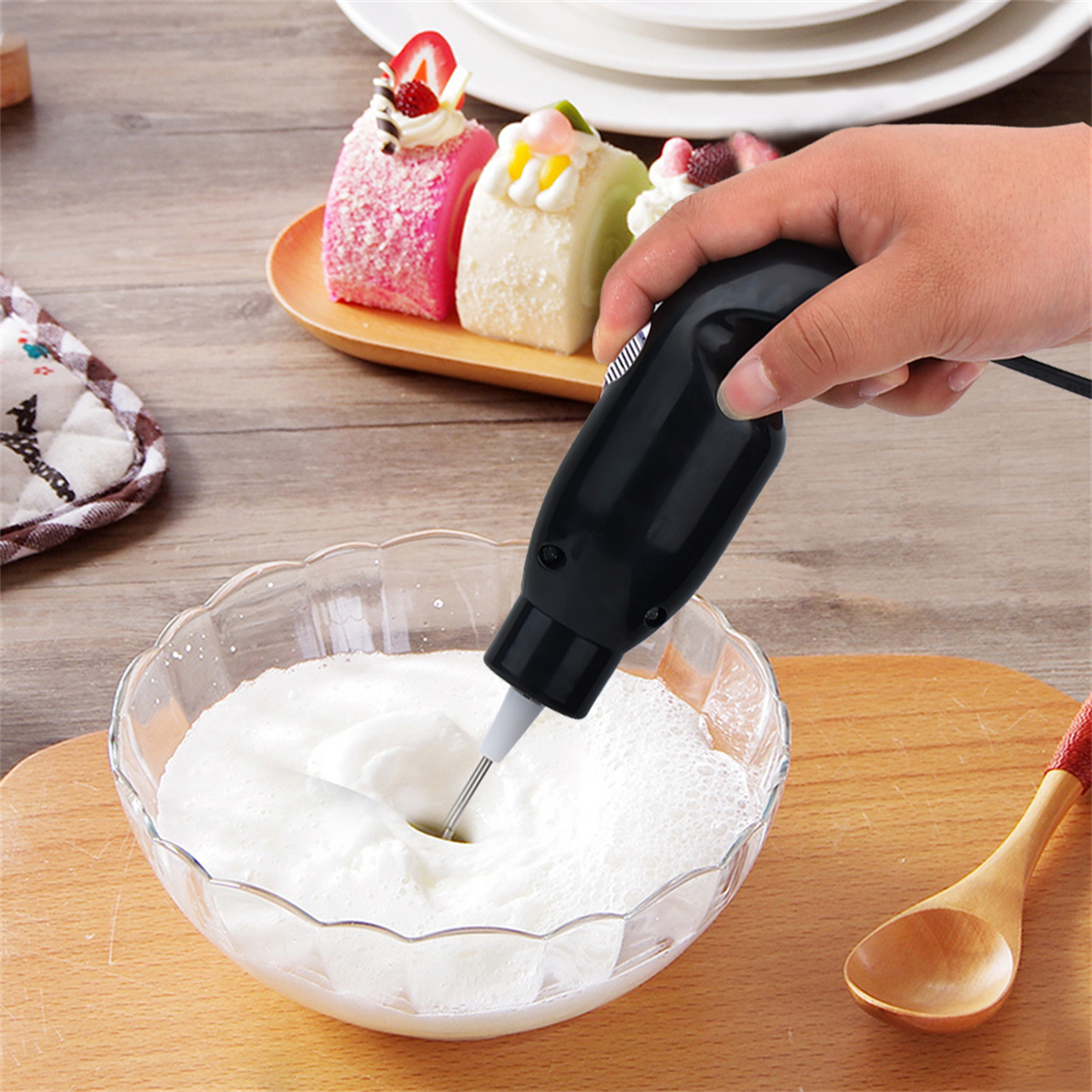 Schwarzer Handmixer, der Schaum in einer Glasschüssel verrührt, drei Kuchen auf einem Teller und ein Holzlöffel.