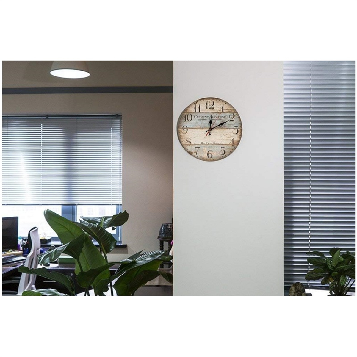 Eine Wanduhr mit römischen Ziffern hängt an einer weißen Wand zwischen Jalousienfenstern.