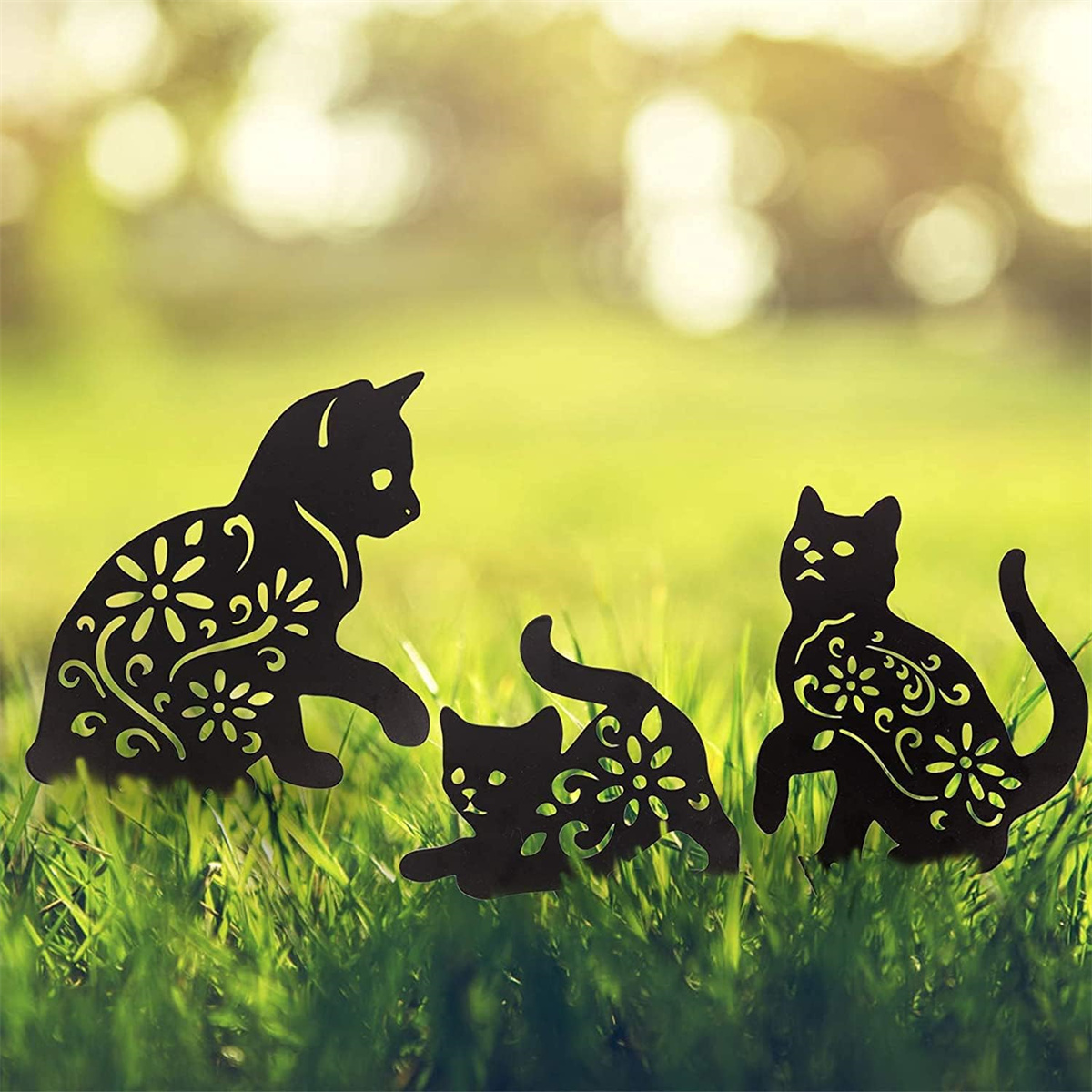 Drei schwarze Katzensilhouetten mit floralen Designs auf Gras im Sonnenlicht.