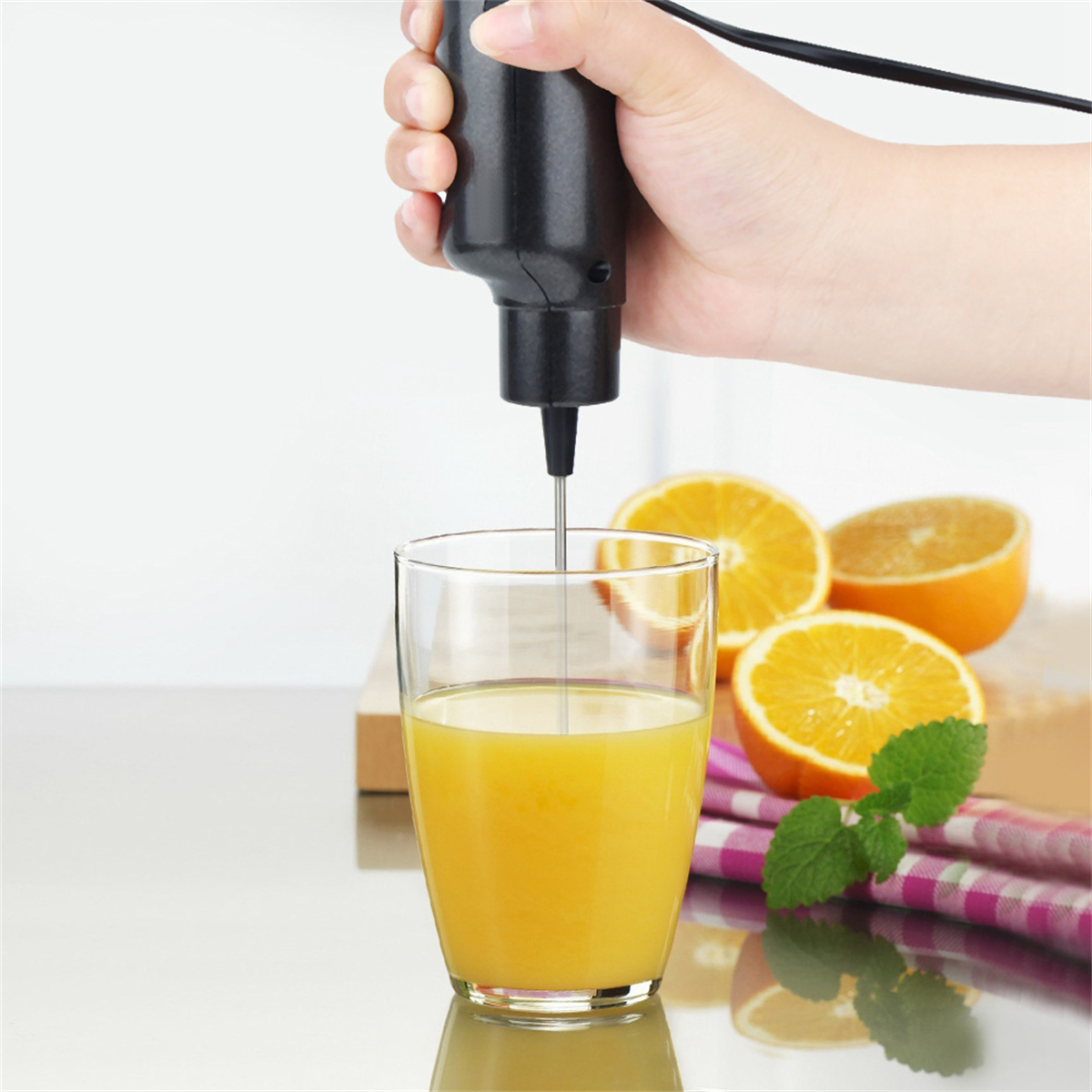 Hand hält Mixer in einem Glas Orangensaft, geschnittene Orangen und Minzblätter auf dem Tisch.