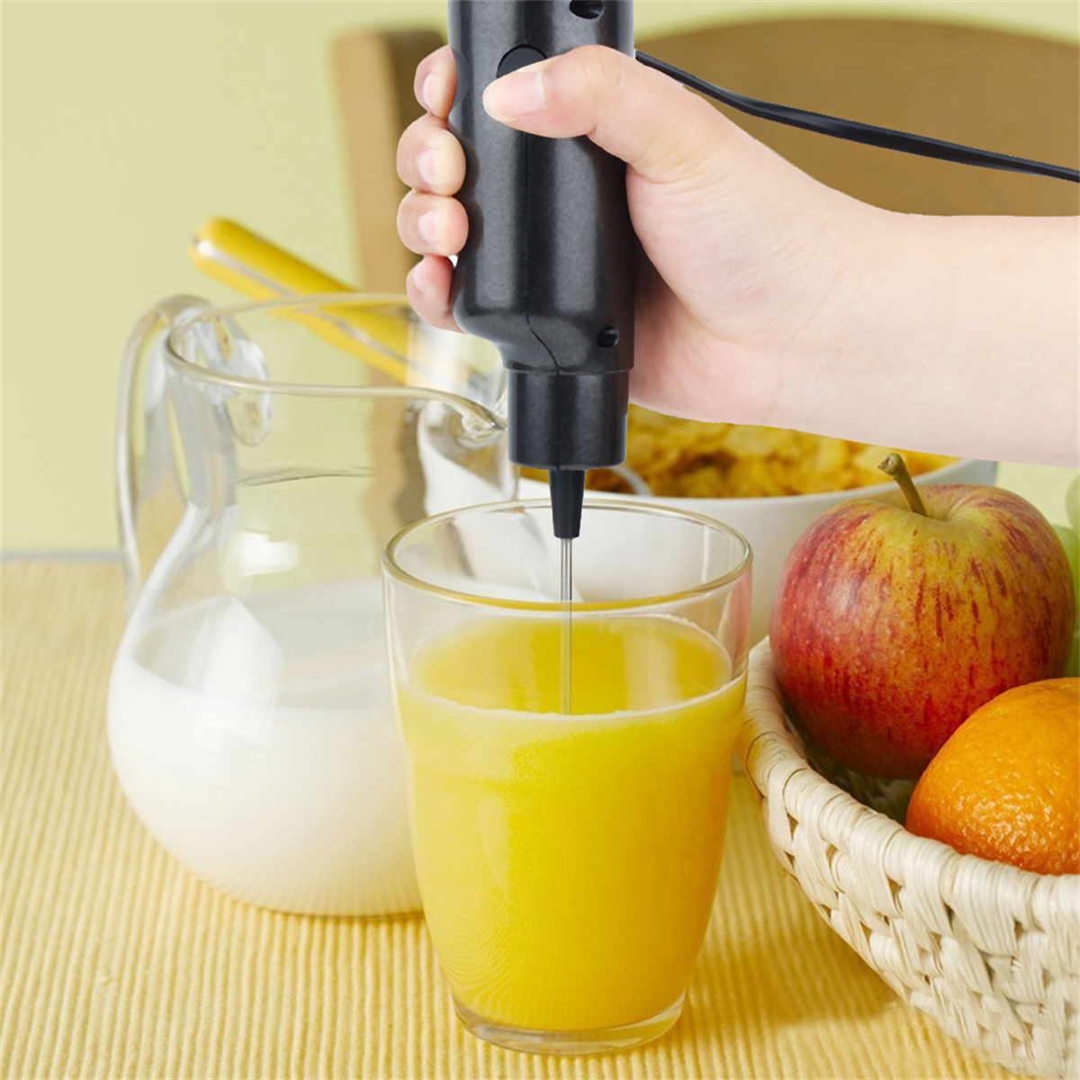 Hand benutzt einen schwarzen Mixer in einem Glas Orangensaft. Milch, Apfel und Orange im Hintergrund.