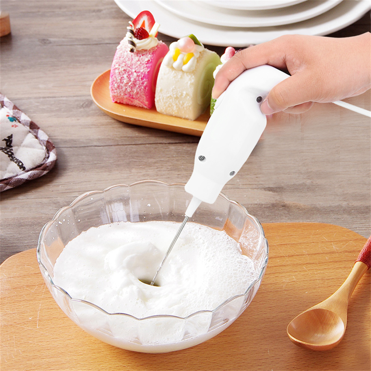 Eine Hand schäumt Milch in einer Glasschüssel auf, Kuchen auf einem Holztablett im Hintergrund.