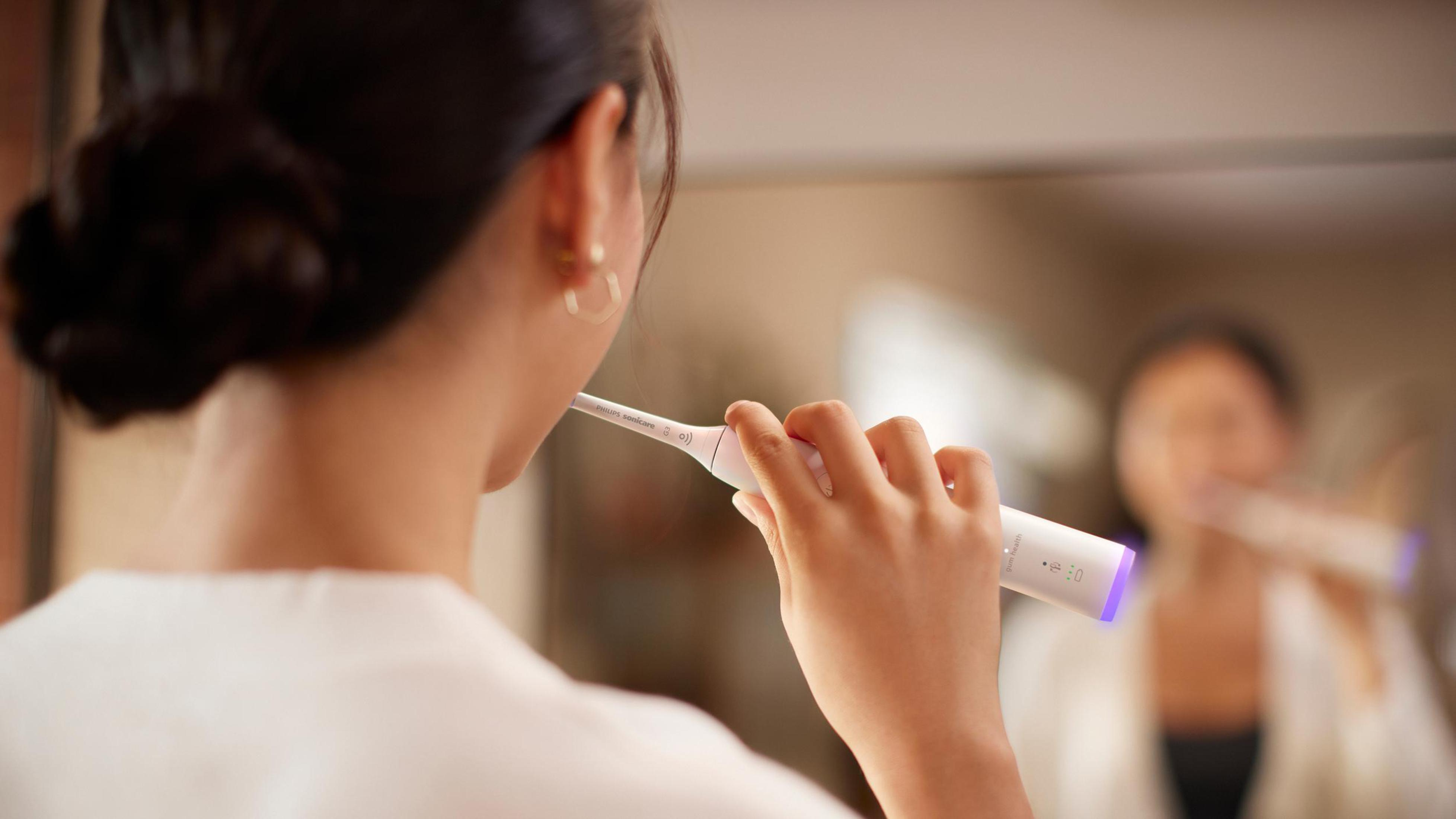 Una mujer cepillándose los dientes frente a un espejo, usando un cepillo de dientes eléctrico.
