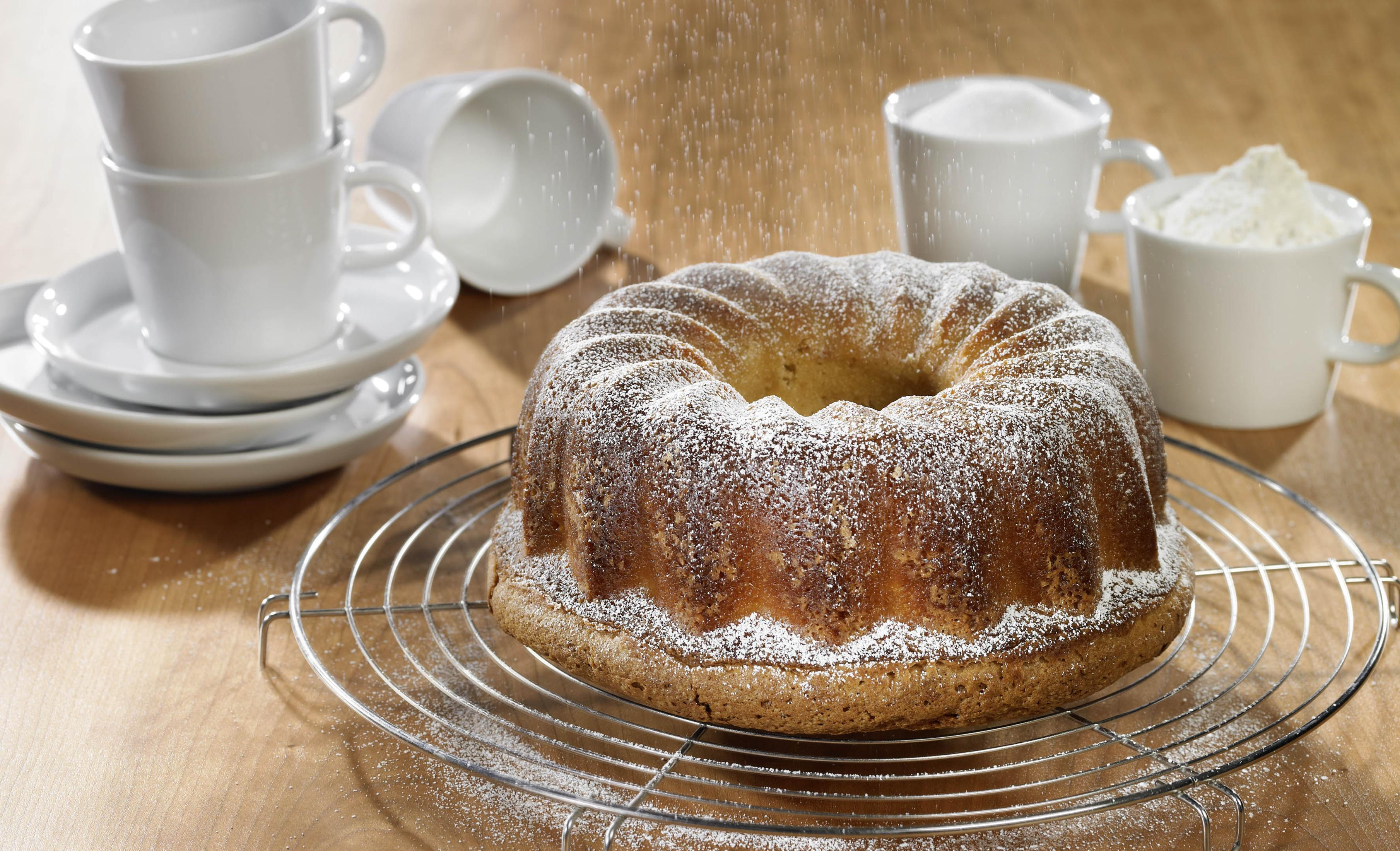 Babka piaskowa posypana cukrem pudrem na metalowej kratce, z kilkoma kubkami i naczyniami dookoła.