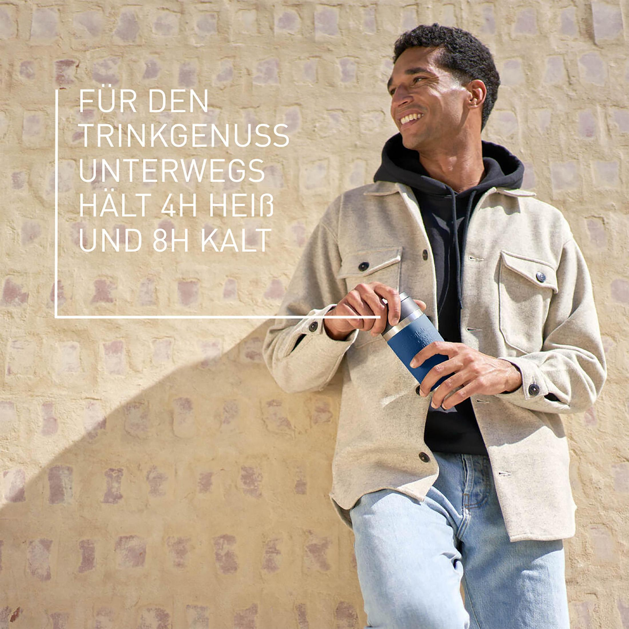 Ein Mann hält eine blaue Thermoskanne vor einer strukturierten Wand.