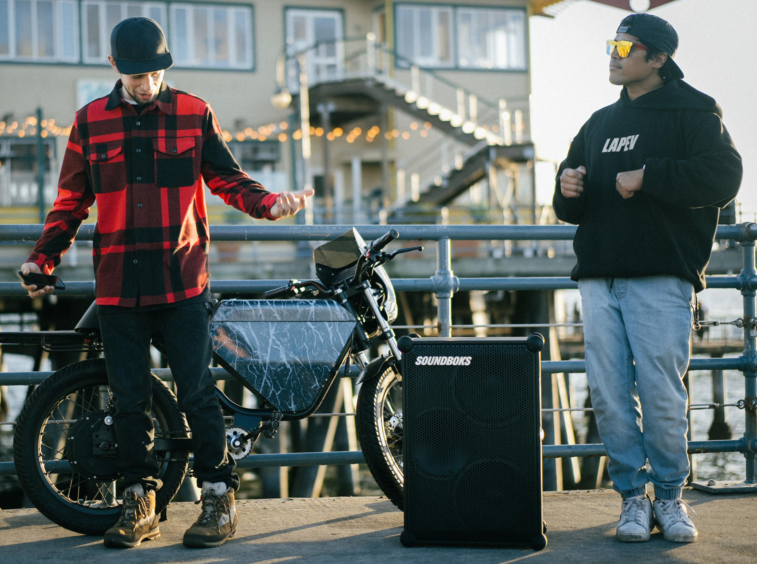 Zwei Männer in der Nähe eines Motorrads und eines Lautsprechers auf einem Pier. Ein Mann gestikuliert, der andere trägt eine Sonnenbrille.