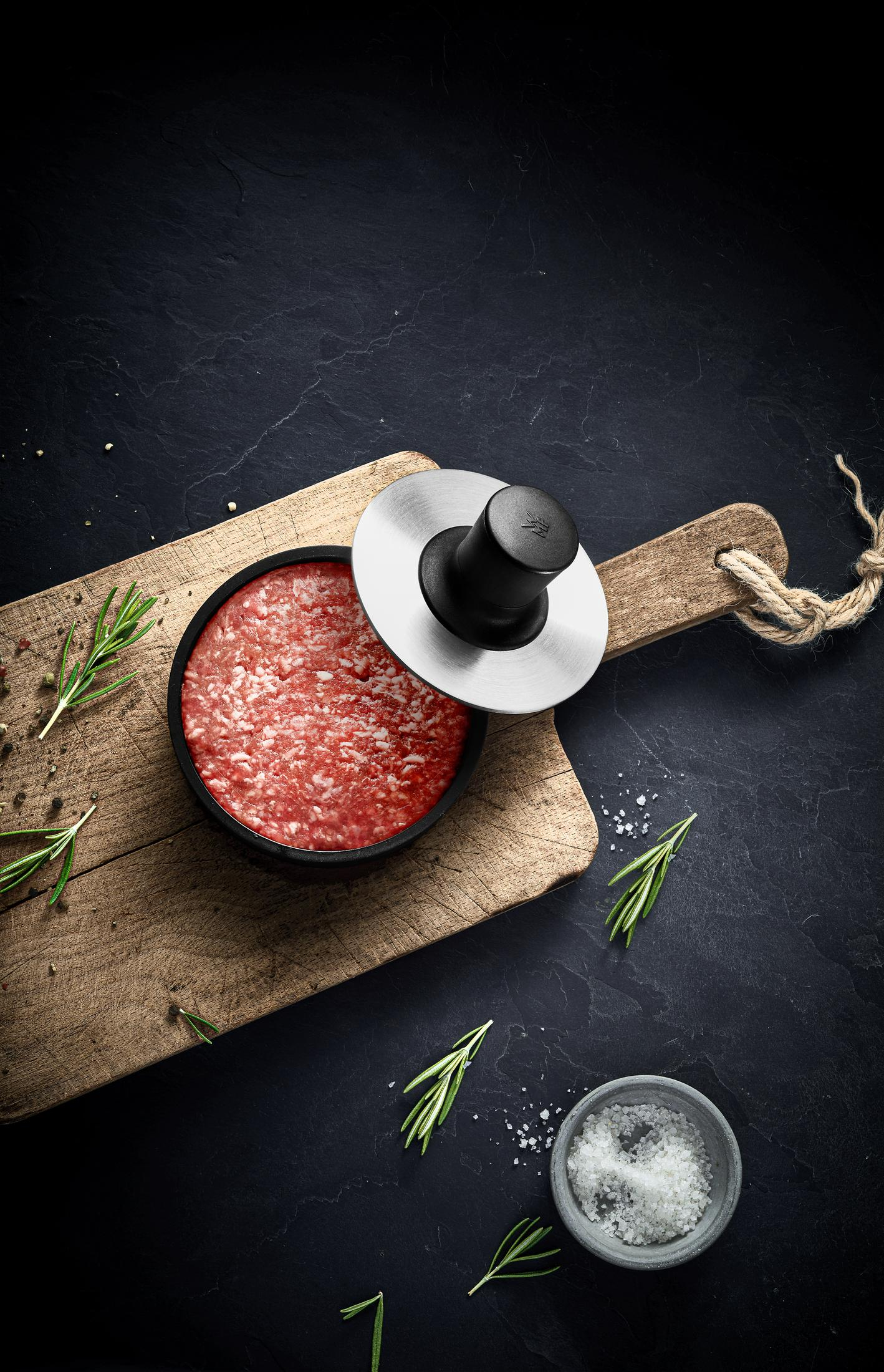 Burgerpresse auf einem Schneidebrett mit Hackfleisch. Rosmarin und Salz liegen daneben.