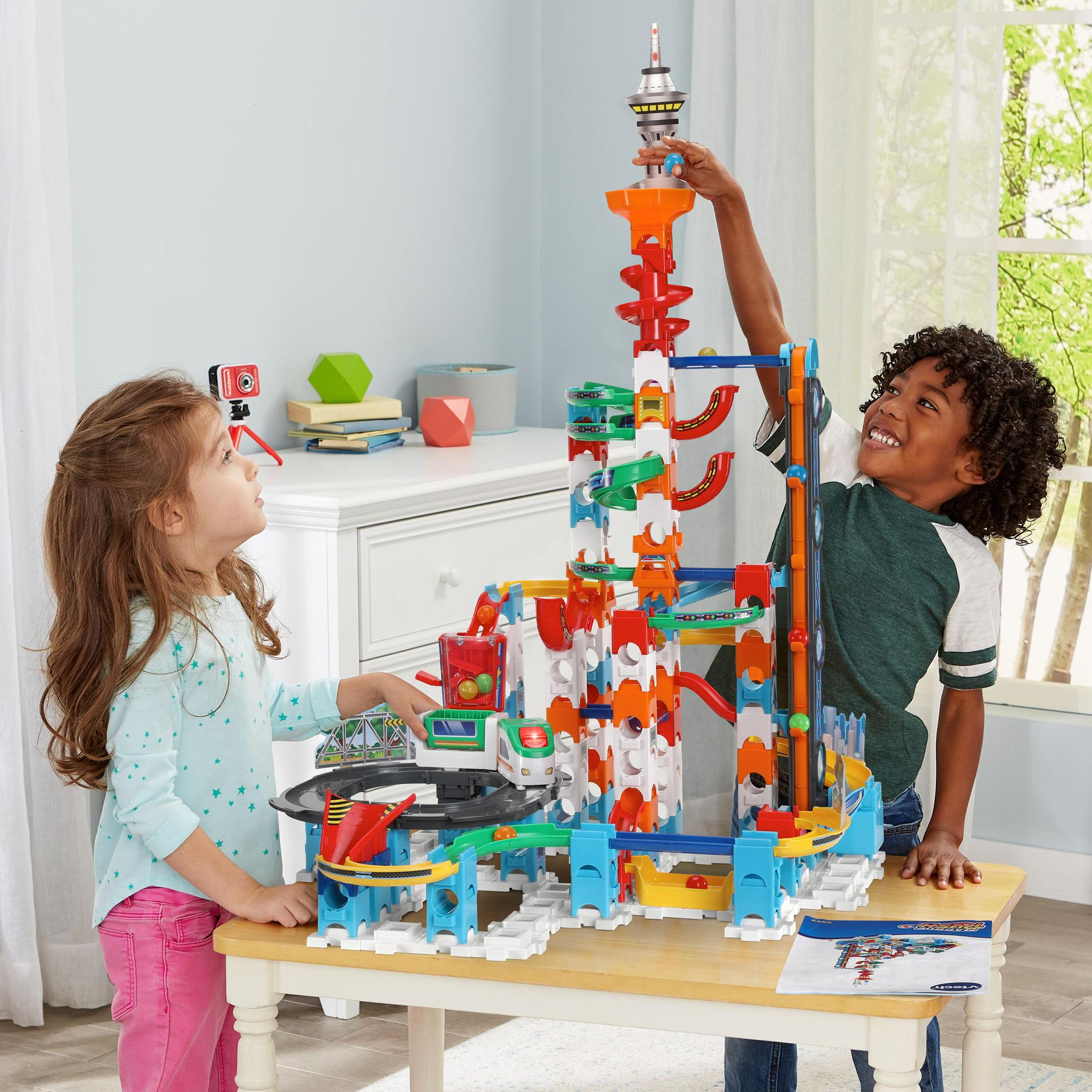 Zwei Kinder spielen mit einem Spielzeug-Konstruktionsset. Das Set ist bunt und steht auf einem Tisch. Im Hintergrund ist ein Zimmer.