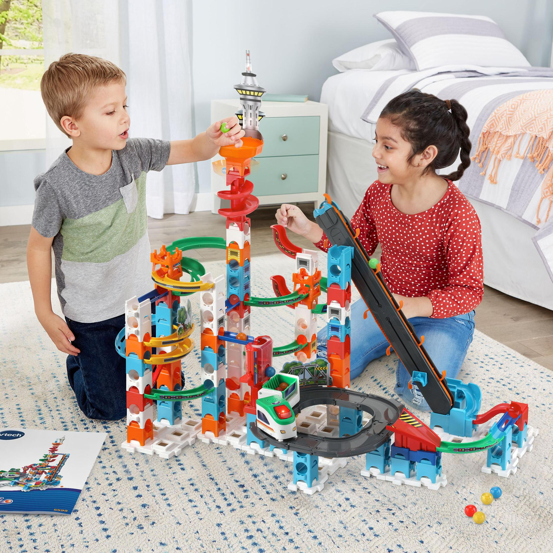 Zwei Kinder spielen mit einem bunten Spielzeugset mit Bahnen, Türmen und einem Fahrzeug auf einem Teppich.