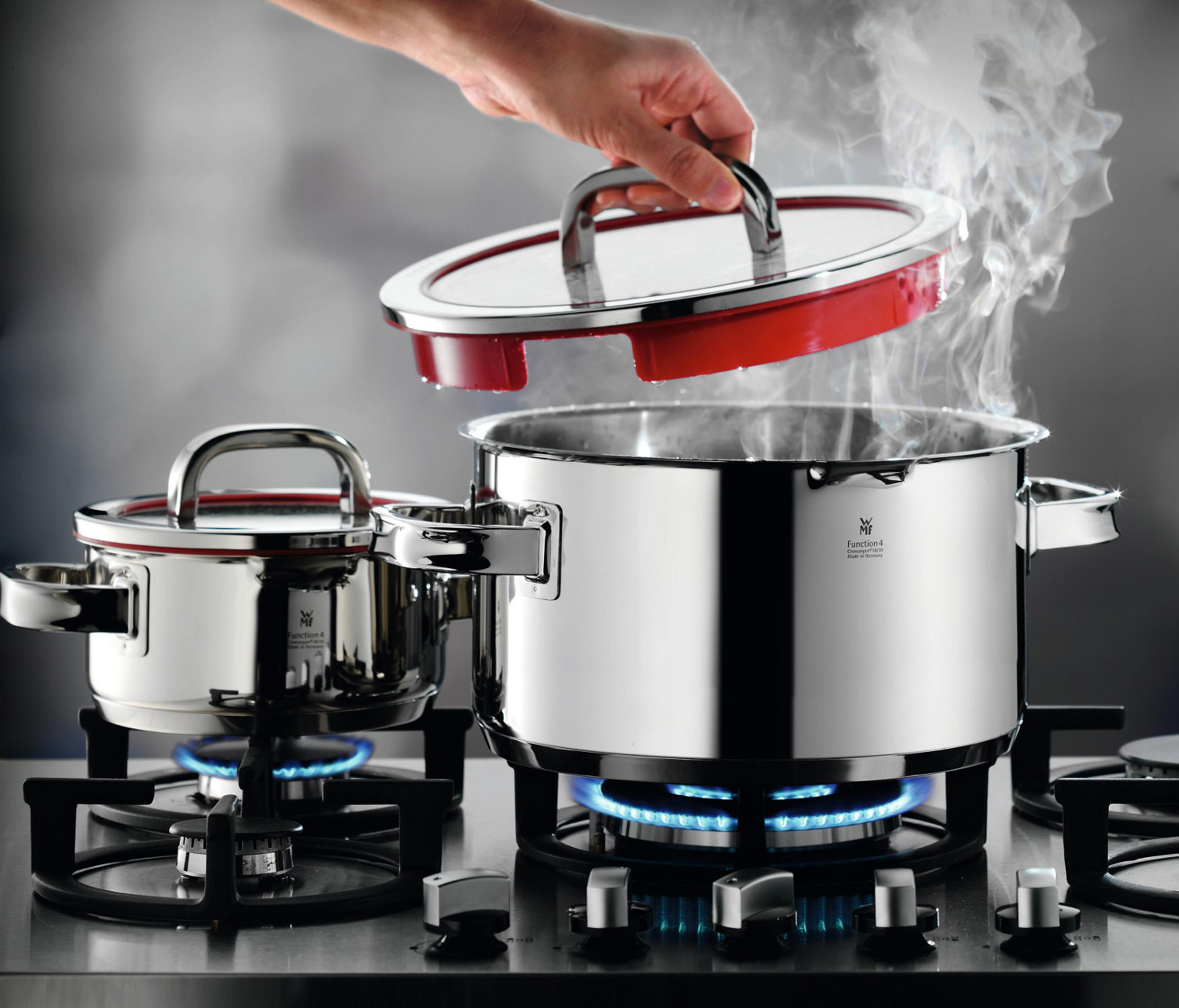 Edelstahl-Kochtopf auf einem Gasherd, eine Hand hebt den Deckel, Dampf steigt auf.