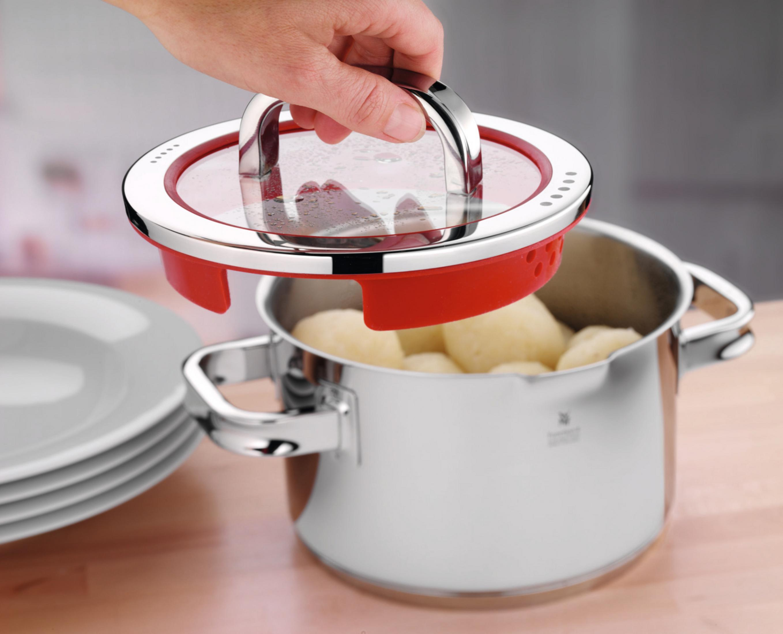 Eine Hand hält einen roten Dampfgarer-Deckel über einem Topf aus Edelstahl, der mit Kartoffeln gefüllt ist.