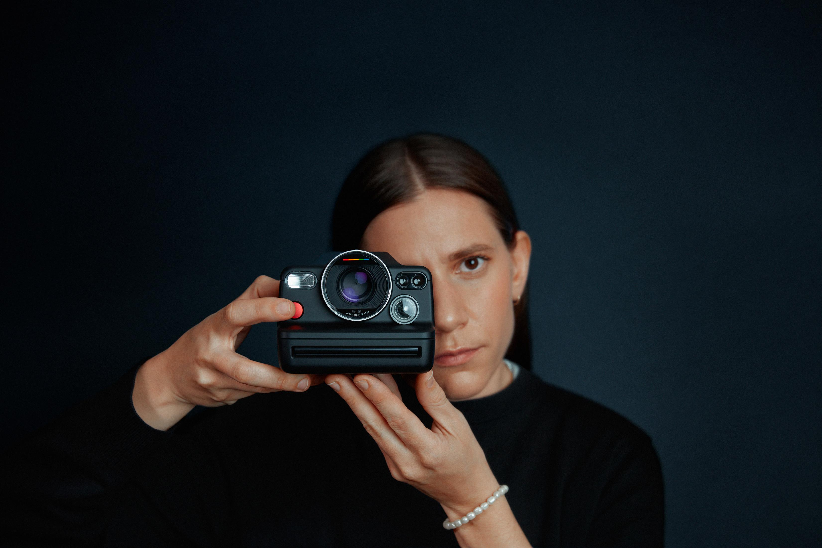 Siyah bir Polaroid fotoğraf makinesi tutan kadın, koyu bir arka plan üzerinde izleyiciye bakıyor.