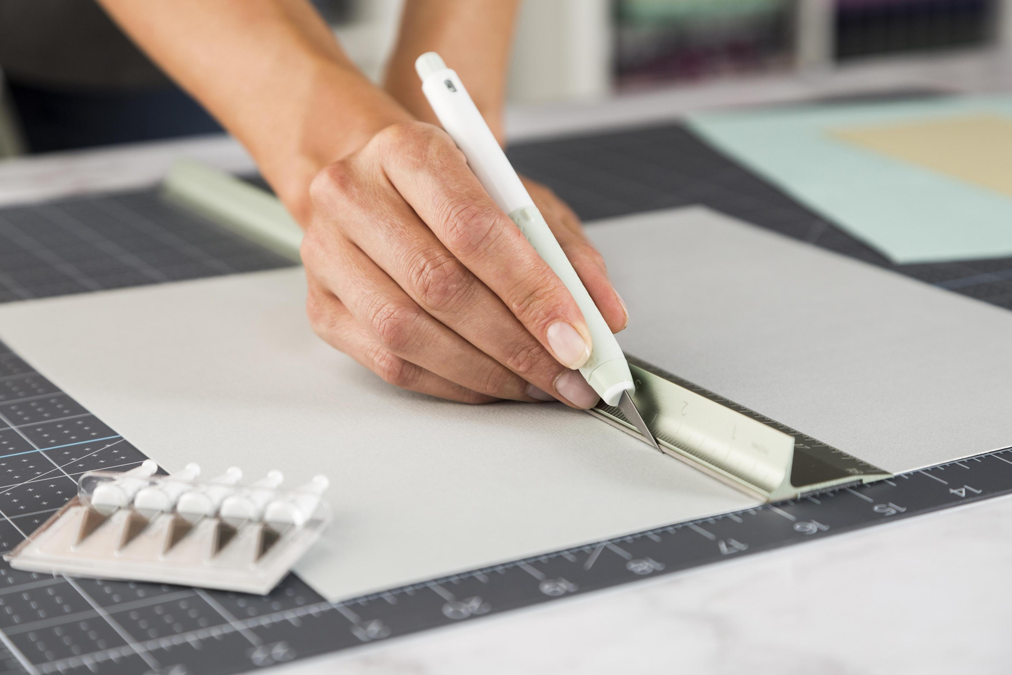 Person schneidet Papier mit einem Bastelmesser und Lineal auf einer Schneidematte. Eine kleine Schale mit Stiften ist zu sehen.