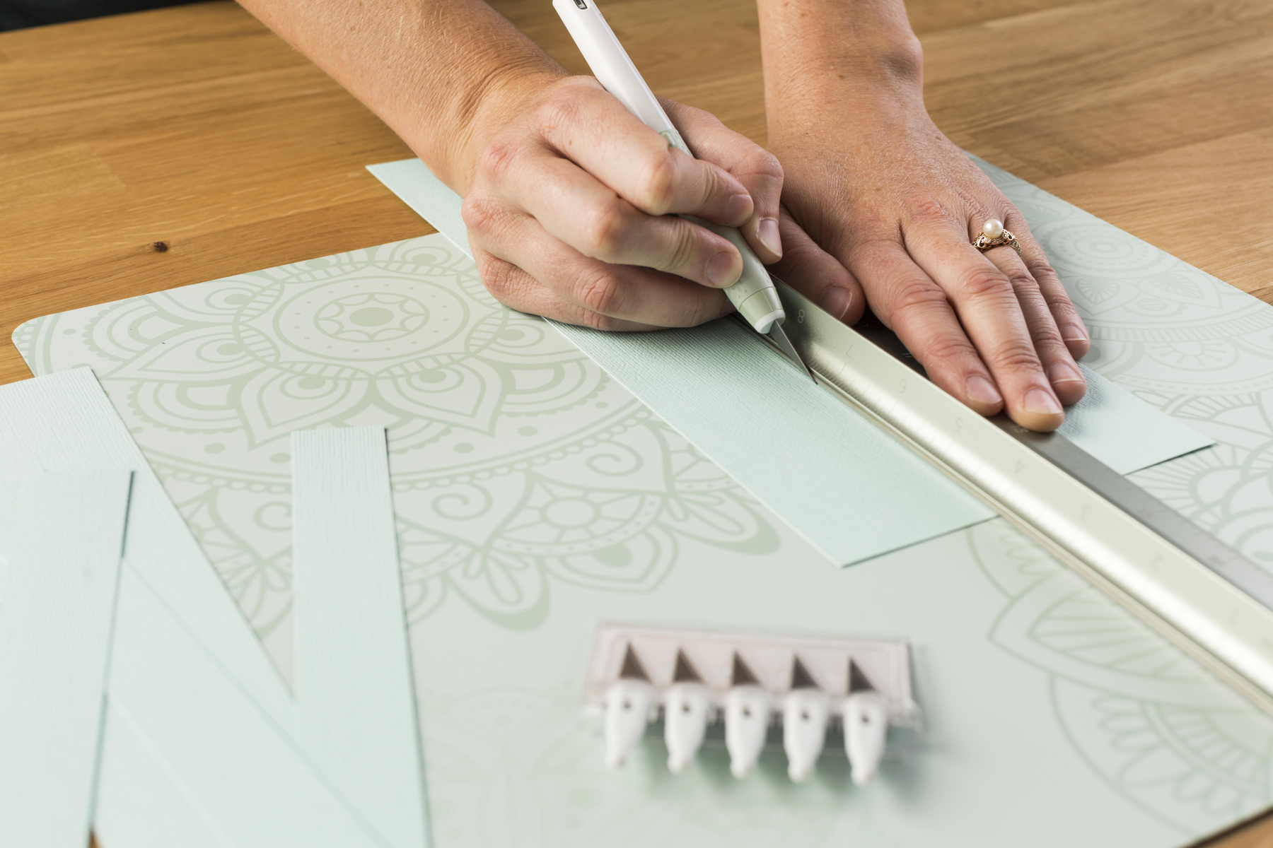 Hände schneiden Papier mit einem Bastelmesser und Lineal auf einer gemusterten Schneidematte.