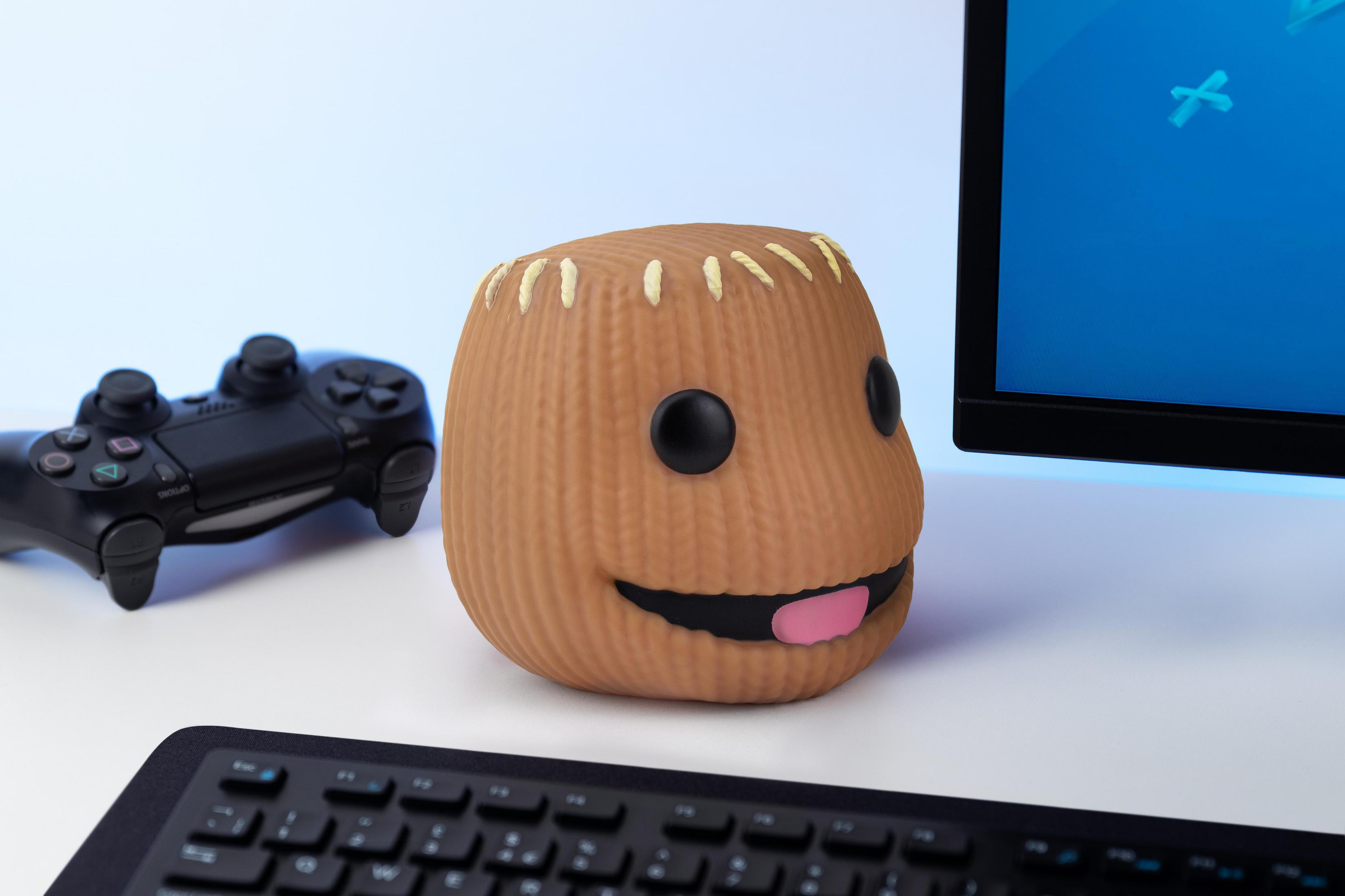 Ein braunes, gestricktes Charakterspielzeug mit schwarzen Augen und einem lächelnden Gesicht, mit einem Gamecontroller.