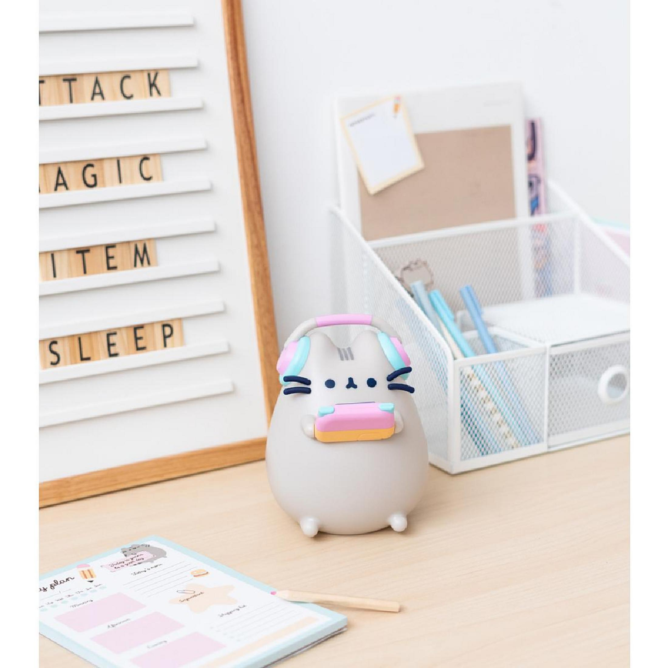 Pusheen-Katzenfigur mit Kopfhörern und einem rosa Gegenstand auf einem Schreibtisch in der Nähe eines Boards.