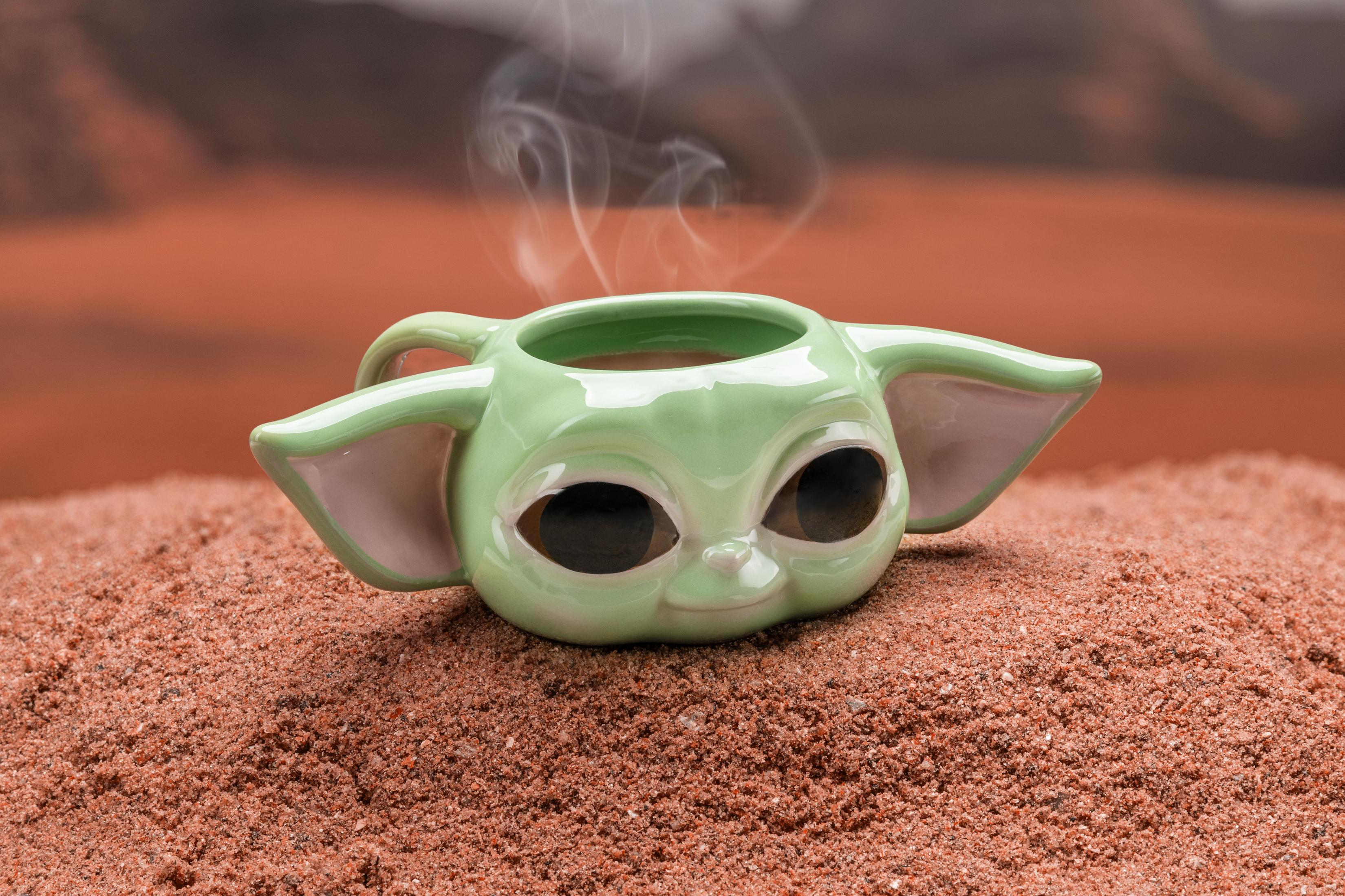 Ein keramischer Grogu (Baby Yoda) Becher mit Dampf, der daraus auf einer sandigen Oberfläche aufsteigt.