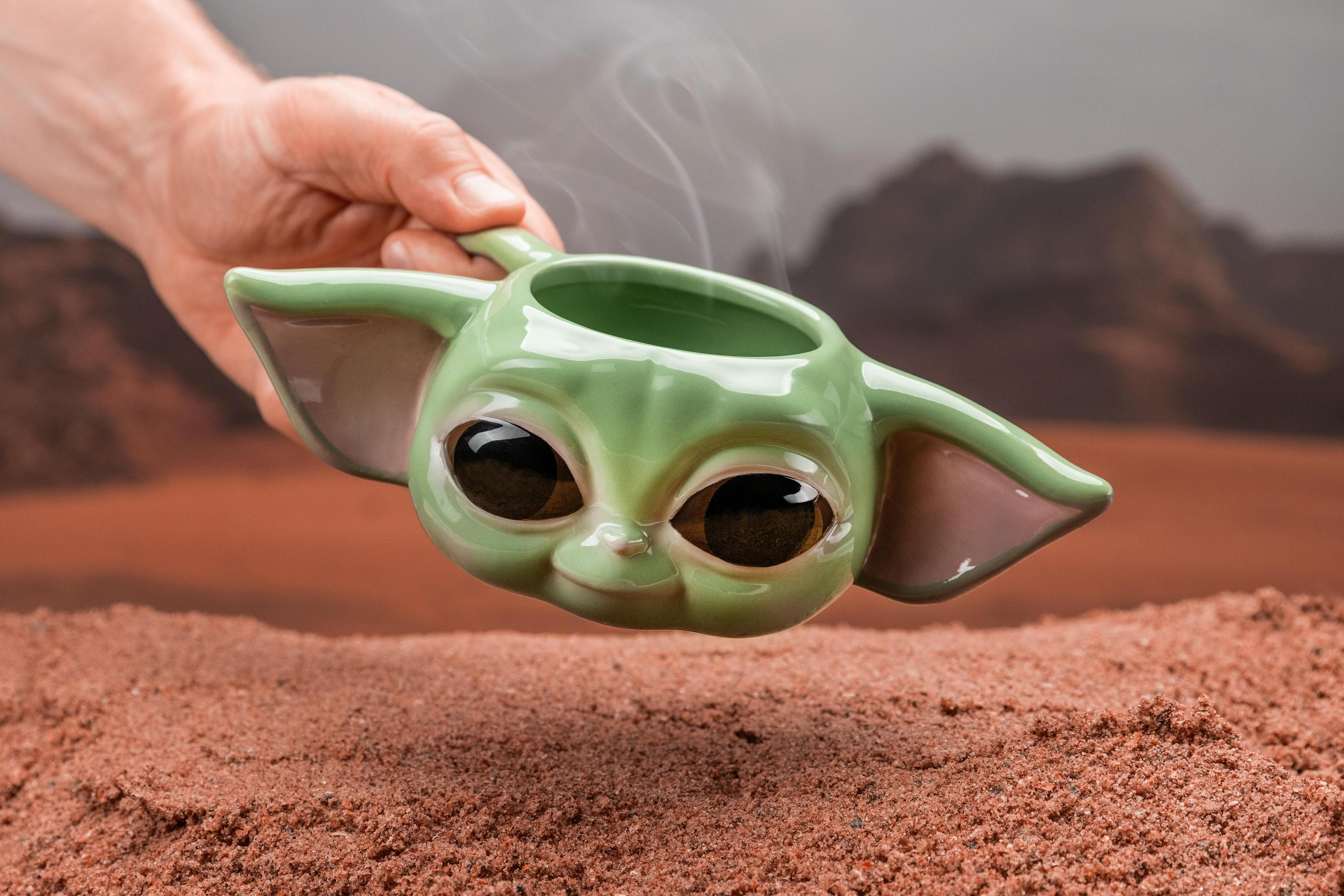 Ein grüner Baby Yoda-Becher mit aufsteigendem Dampf, gehalten von einer Hand vor verschwommenem Hintergrund.