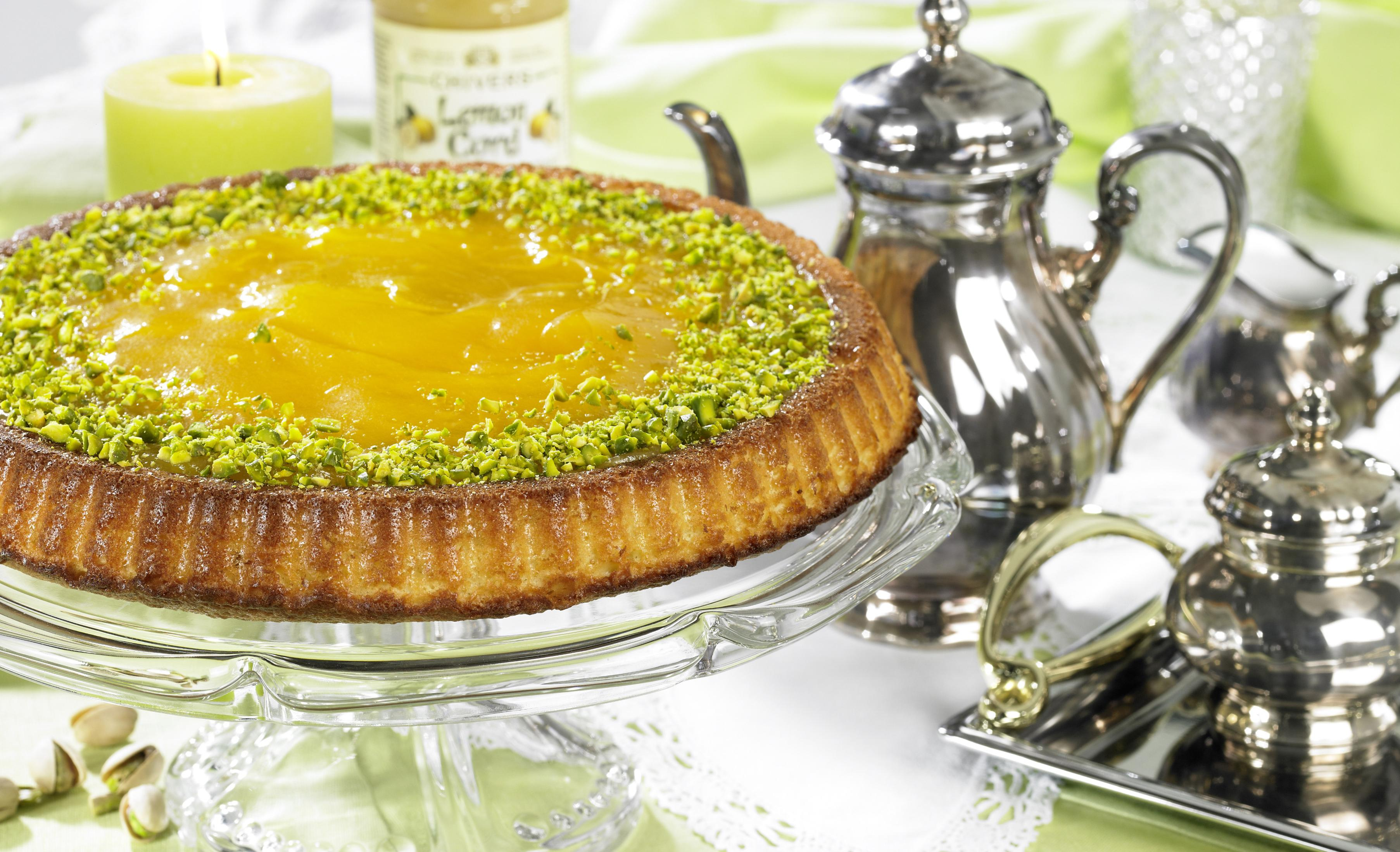 Tarta cytrynowa na szklanej podstawie, z pistacjami, świecą i srebrnym serwisem do herbaty.