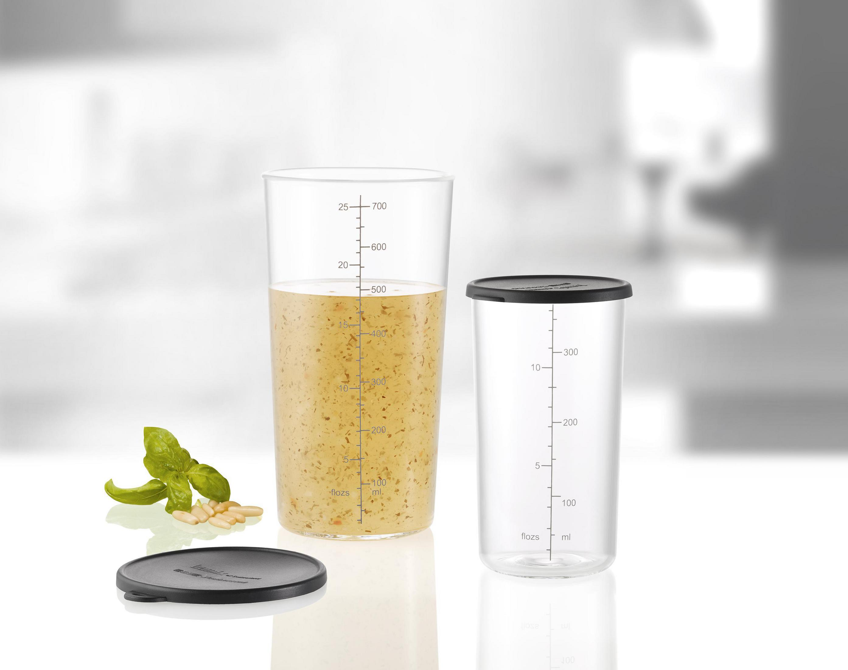Klarer Glaskrug mit Pesto, neben einem kleineren Behälter, mit Deckel und etwas Basilikum.
