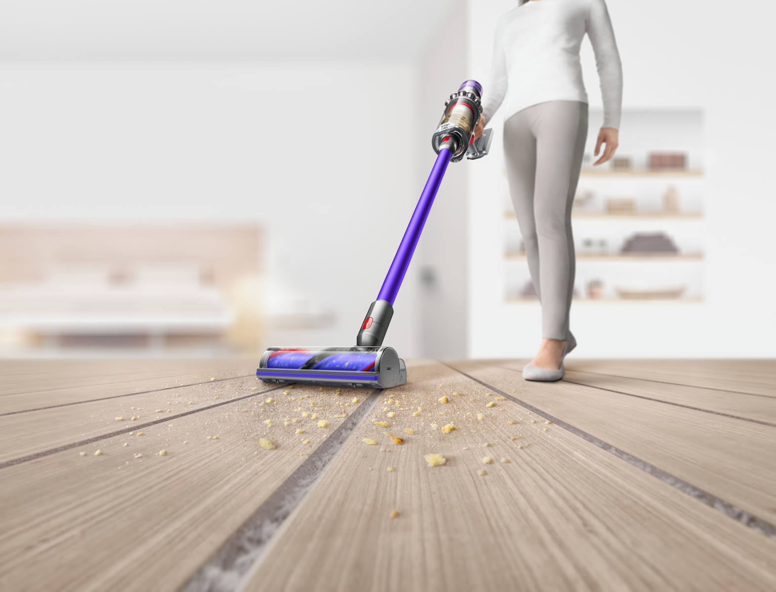Eine Person saugt einen Holzboden mit einem lila-silbernen Dyson-Staubsauger. Krümel sind verstreut.