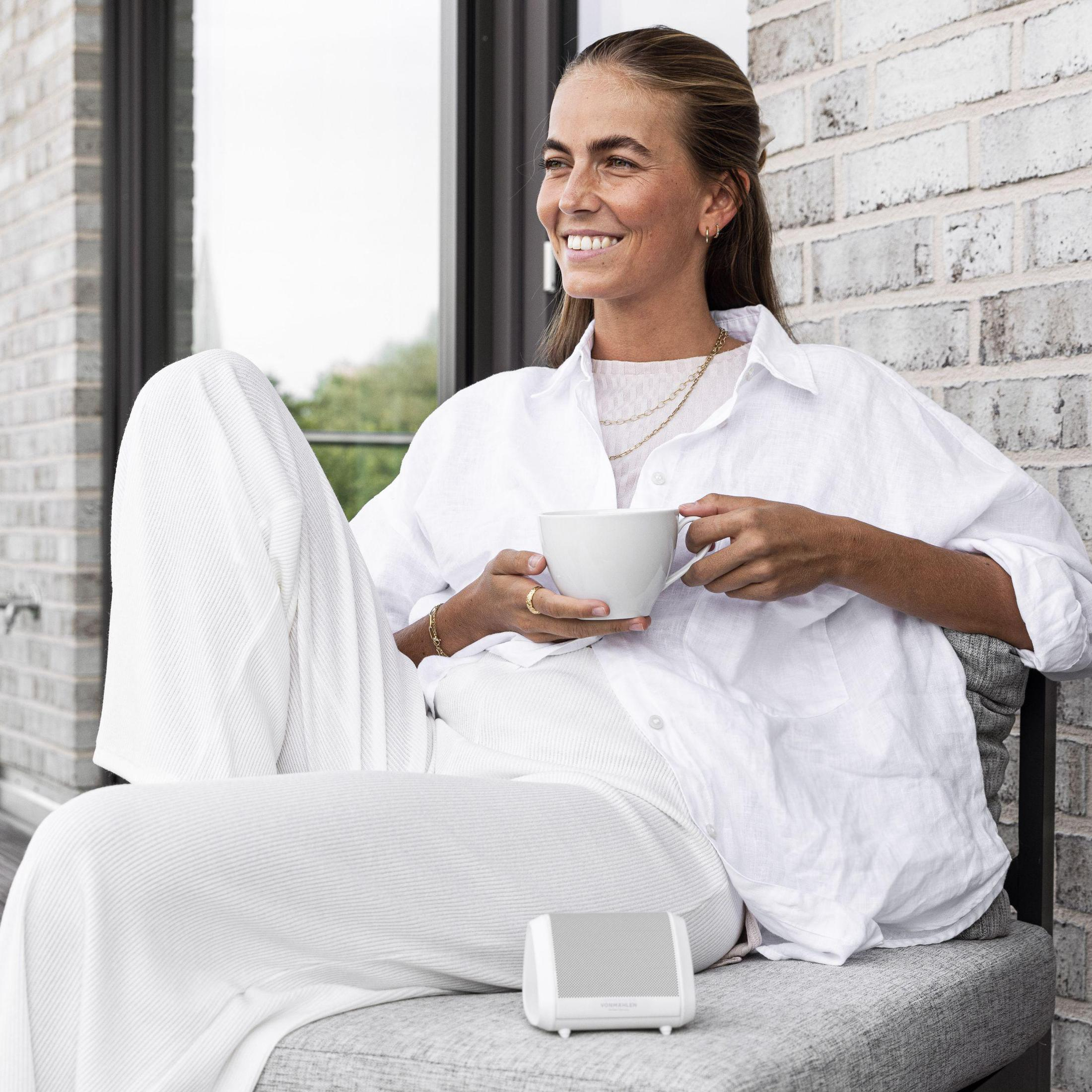 Eine Frau mit einer weißen Tasse sitzt draußen auf einer grauen Bank. Sie lächelt, mit einem weißen Lautsprecher neben sich.