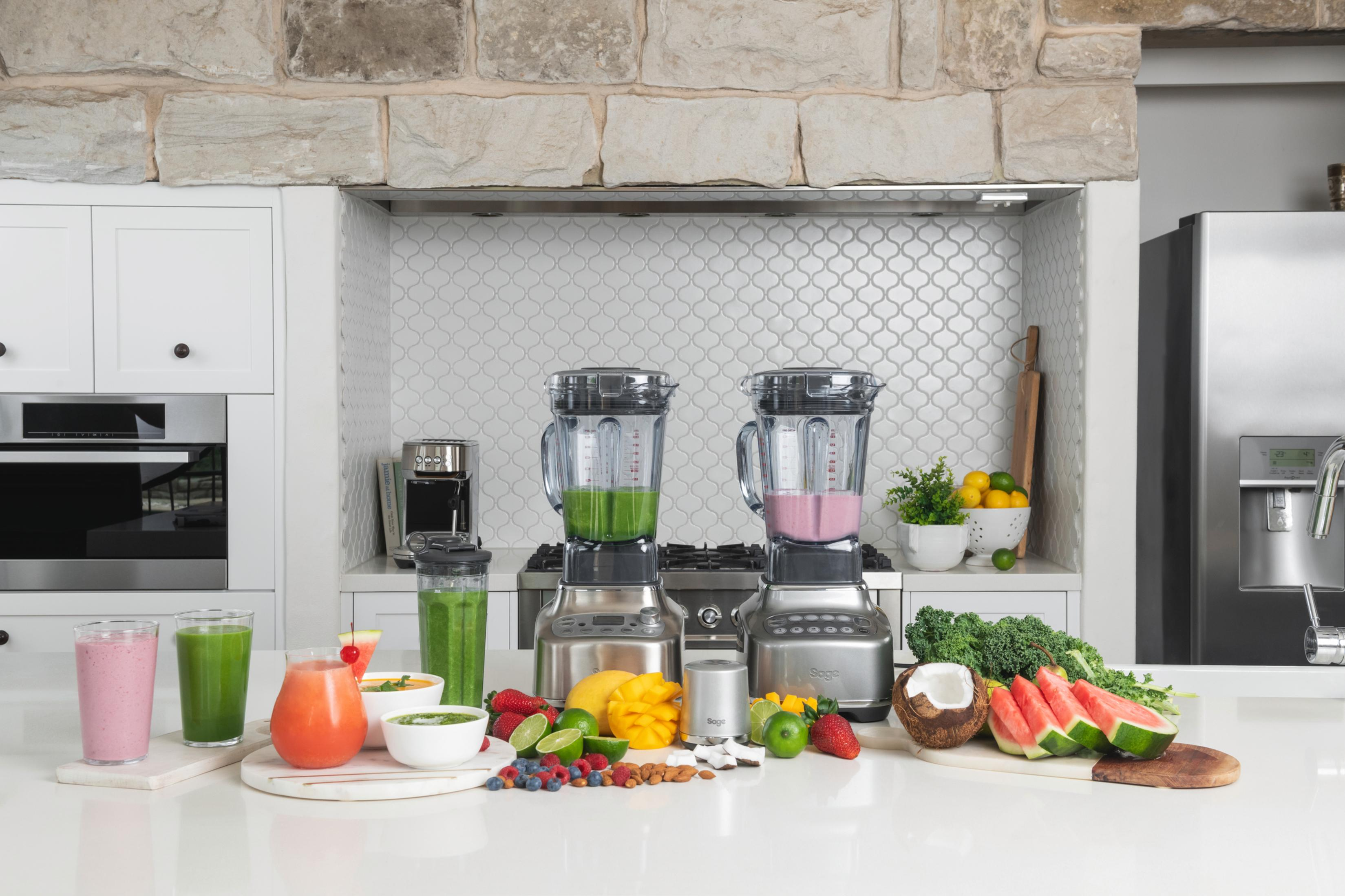 Scena kuchenna z dwoma blenderami, owocami i składnikami na białym blacie. Kuchnia jest biała i szara.