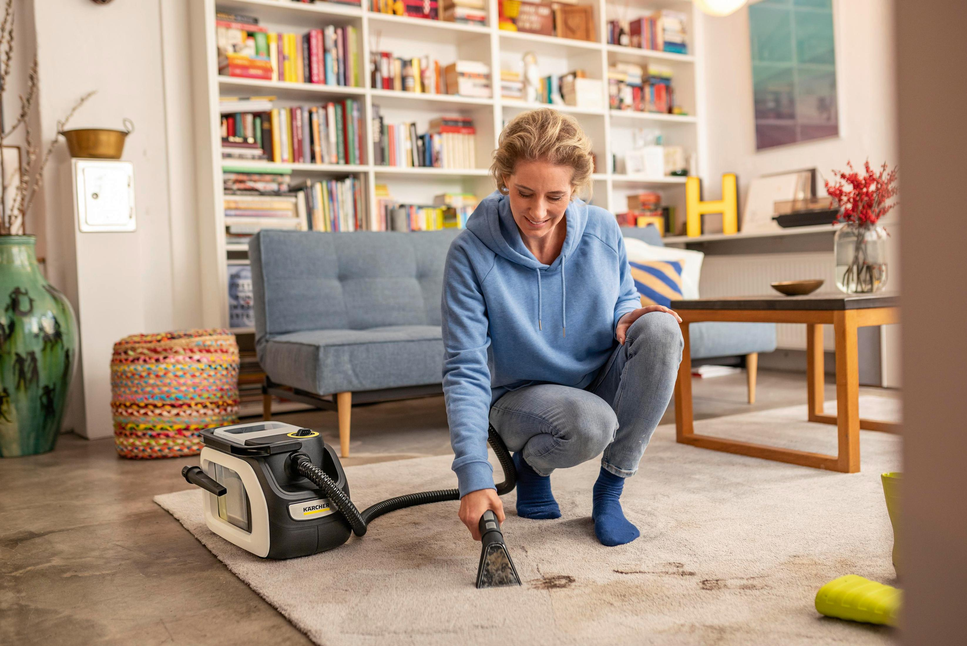Frau reinigt Teppich mit Staubsauger. Das Zimmer hat Couch, Bücherregal und Teppich.