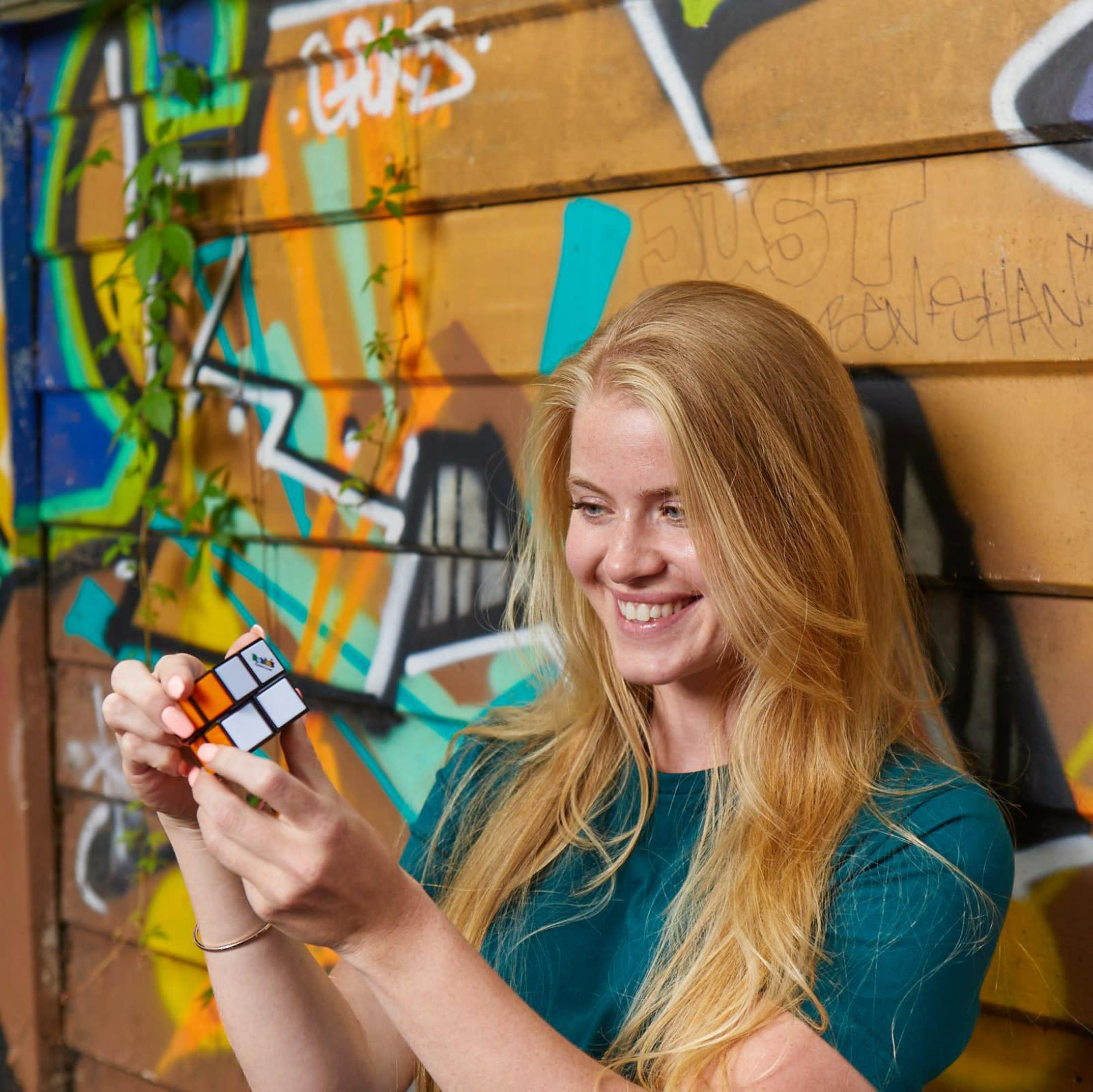 Donna bionda che tiene un cubo di Rubik, sorridendo. Lo sfondo è un muro coperto di graffiti e piante verdi.