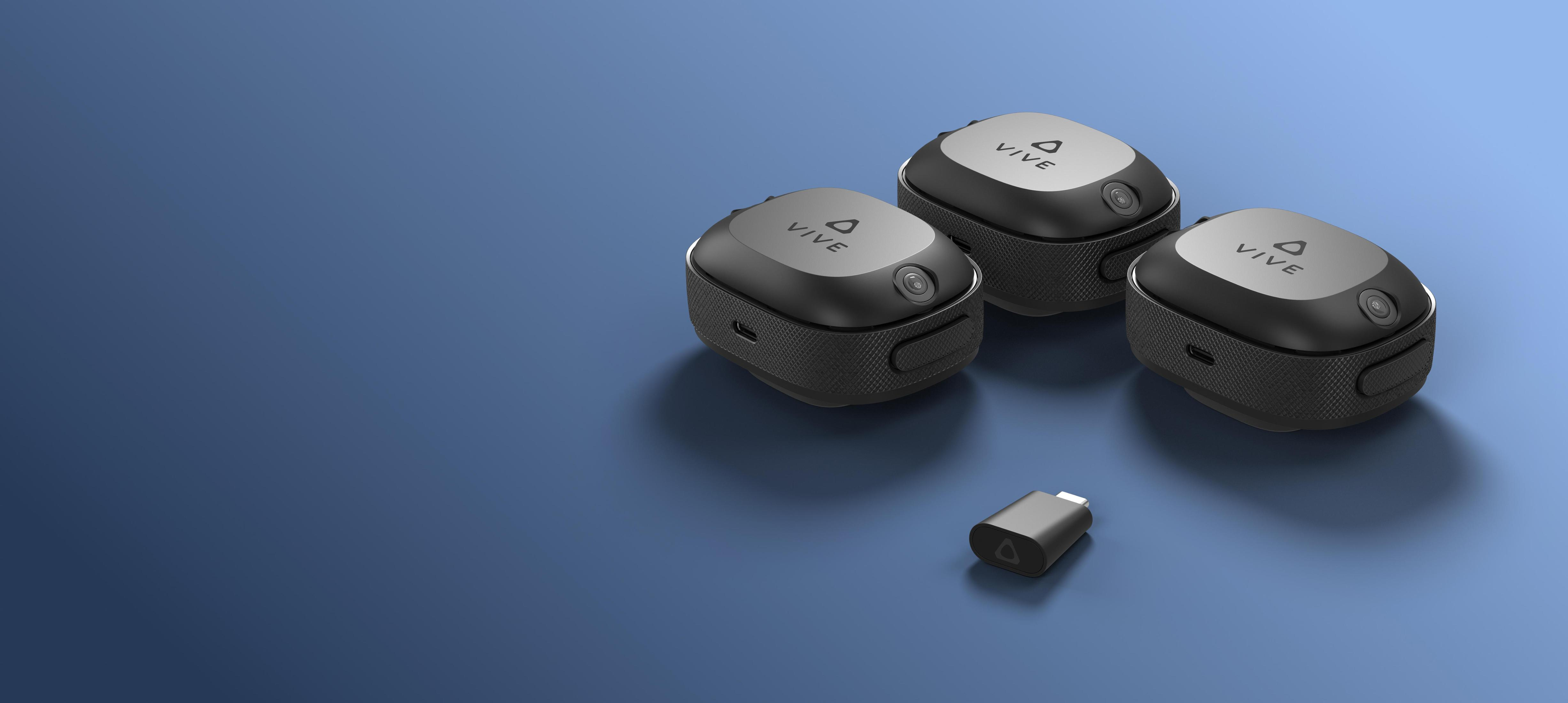 Drei schwarze VR-Tracking-Geräte und ein kleiner USB-Dongle auf blauer Oberfläche.