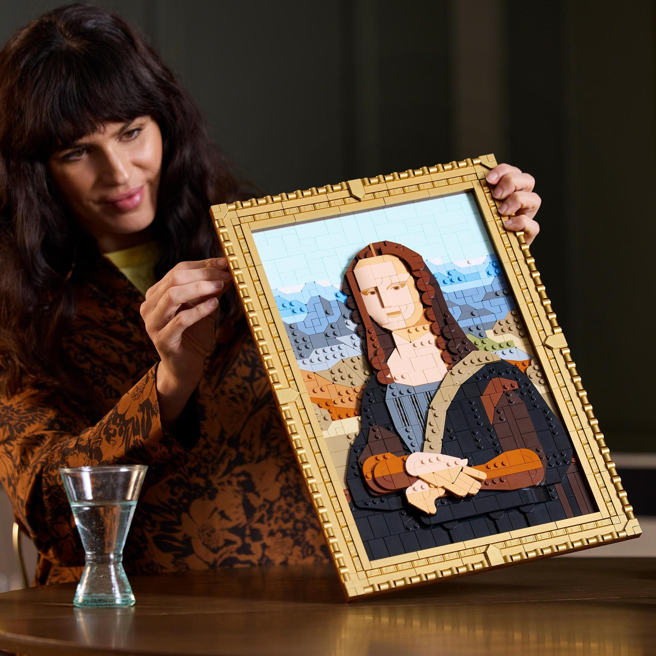 Kobieta trzymająca oprawioną LEGO Mona Lisę. Tło jest ciemne z szklanką wody po lewej.