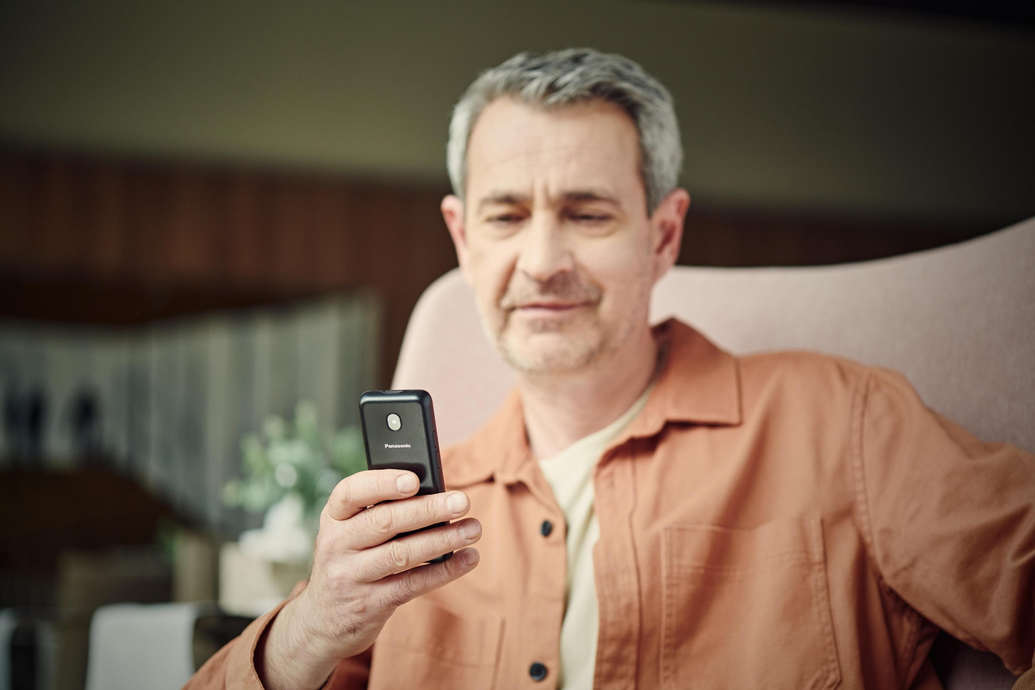 Mann hält Smartphone, schaut drauf. Oranges Hemd, graue Haare. Verschwommener Hintergrund.
