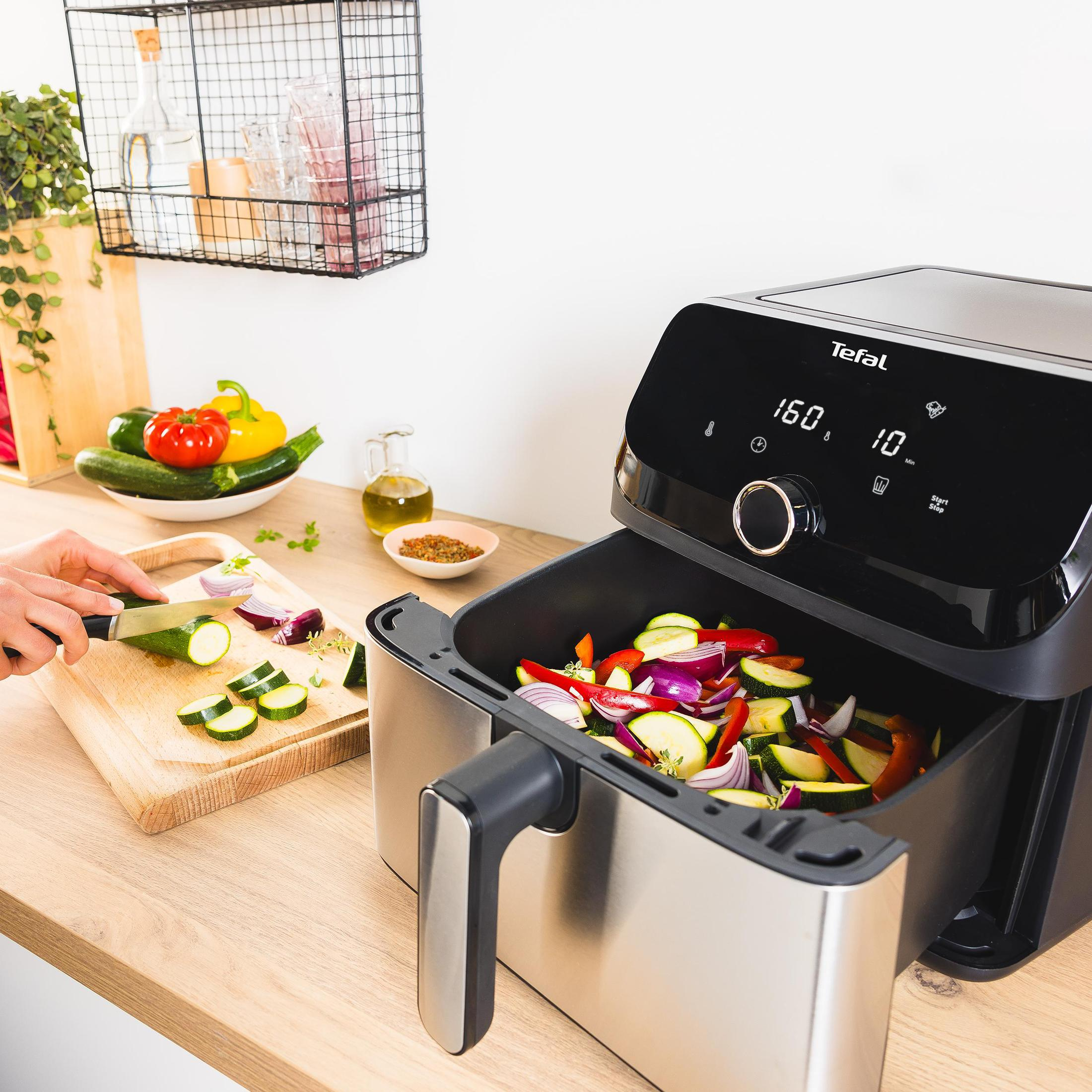 Frytownica powietrzna z warzywami. Osoba krojąca warzywa. Drewniany blat z warzywami w tle.