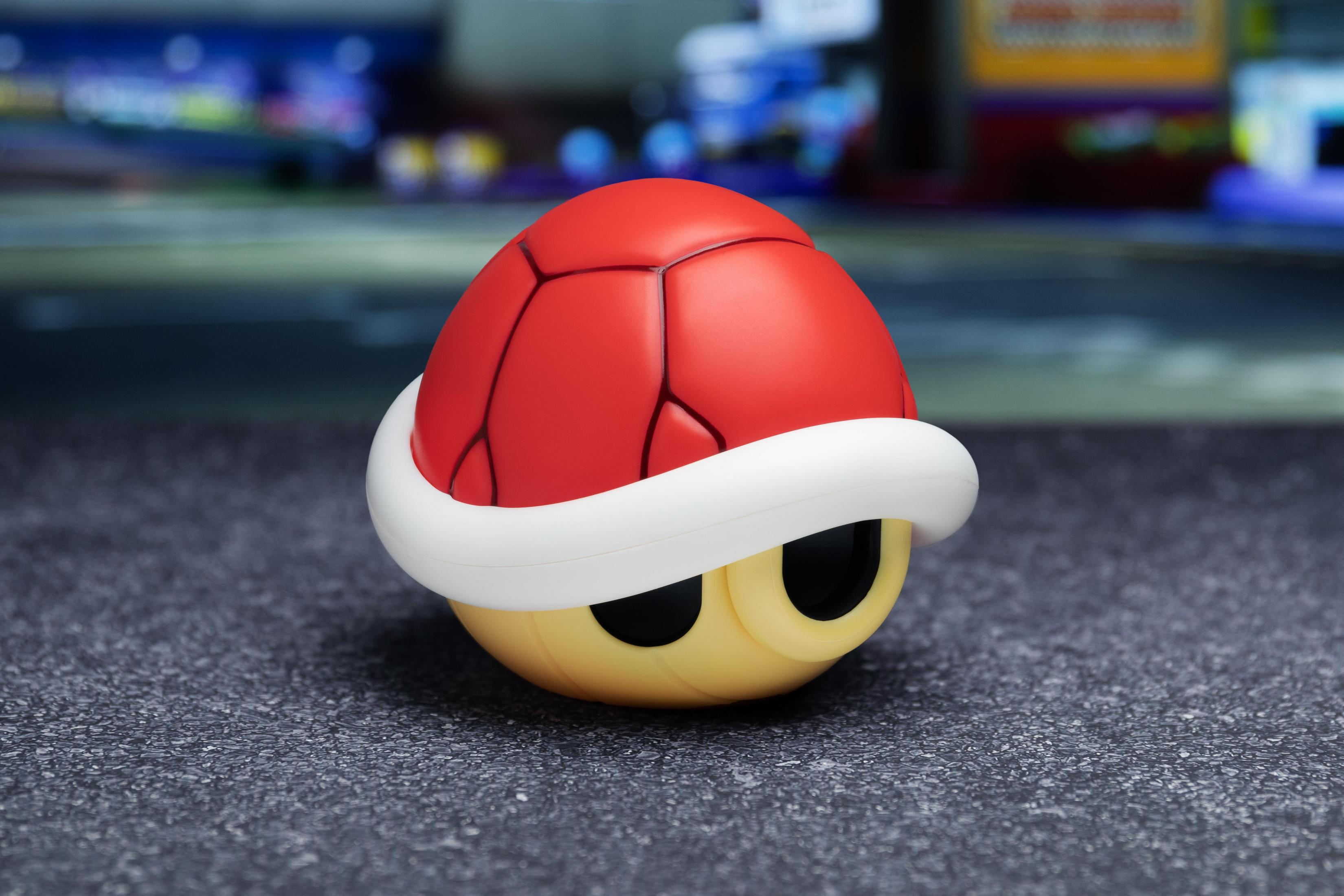 Ein rotes Koopa-Muschelspielzeug mit weißer Umrandung auf grauem Untergrund, verschwommener Stadt.