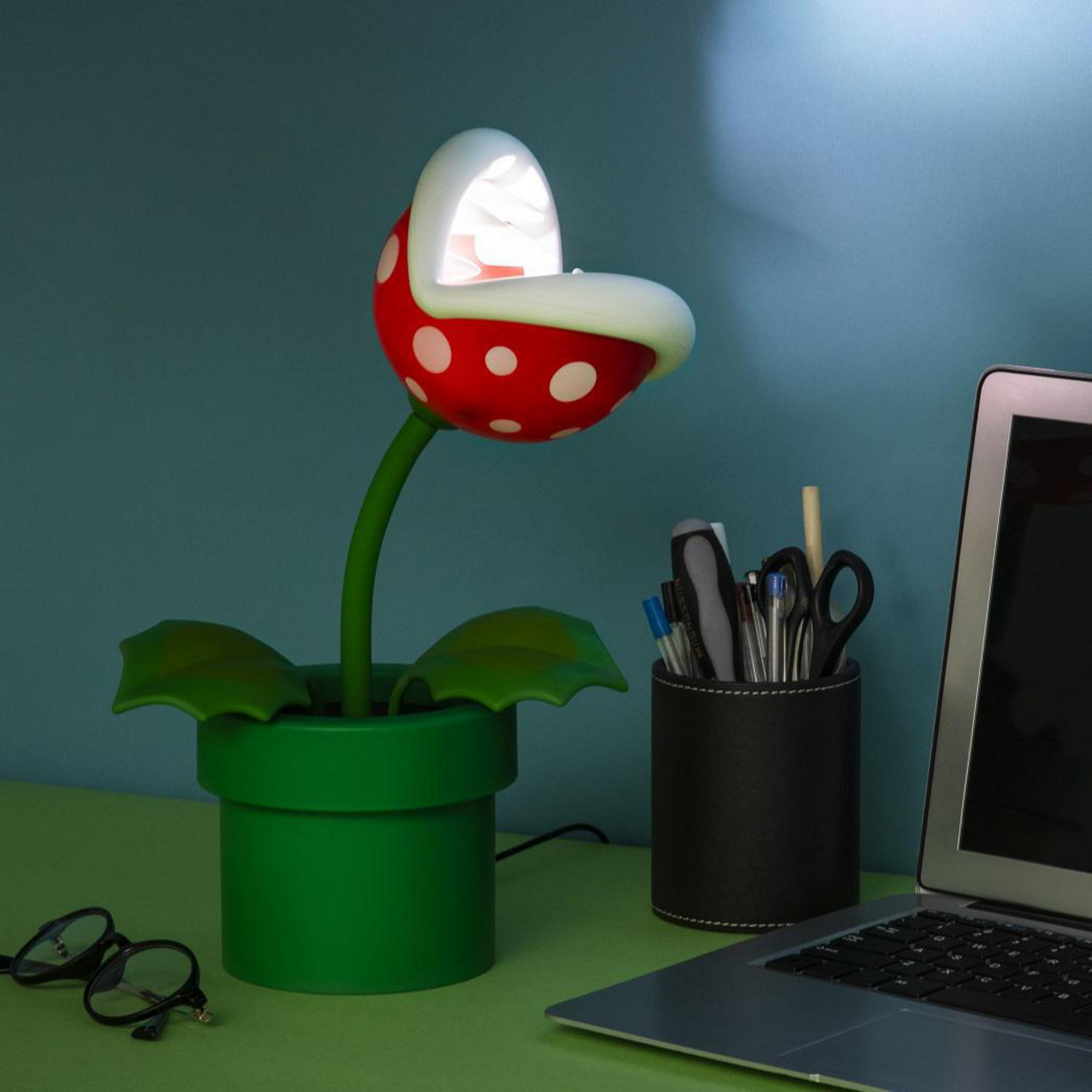 Lampa Piranha Plant z Mario na biurku. Zielony garnek, czerwona roślina z białymi kropkami. Czarny pojemnik na długopisy i laptop.