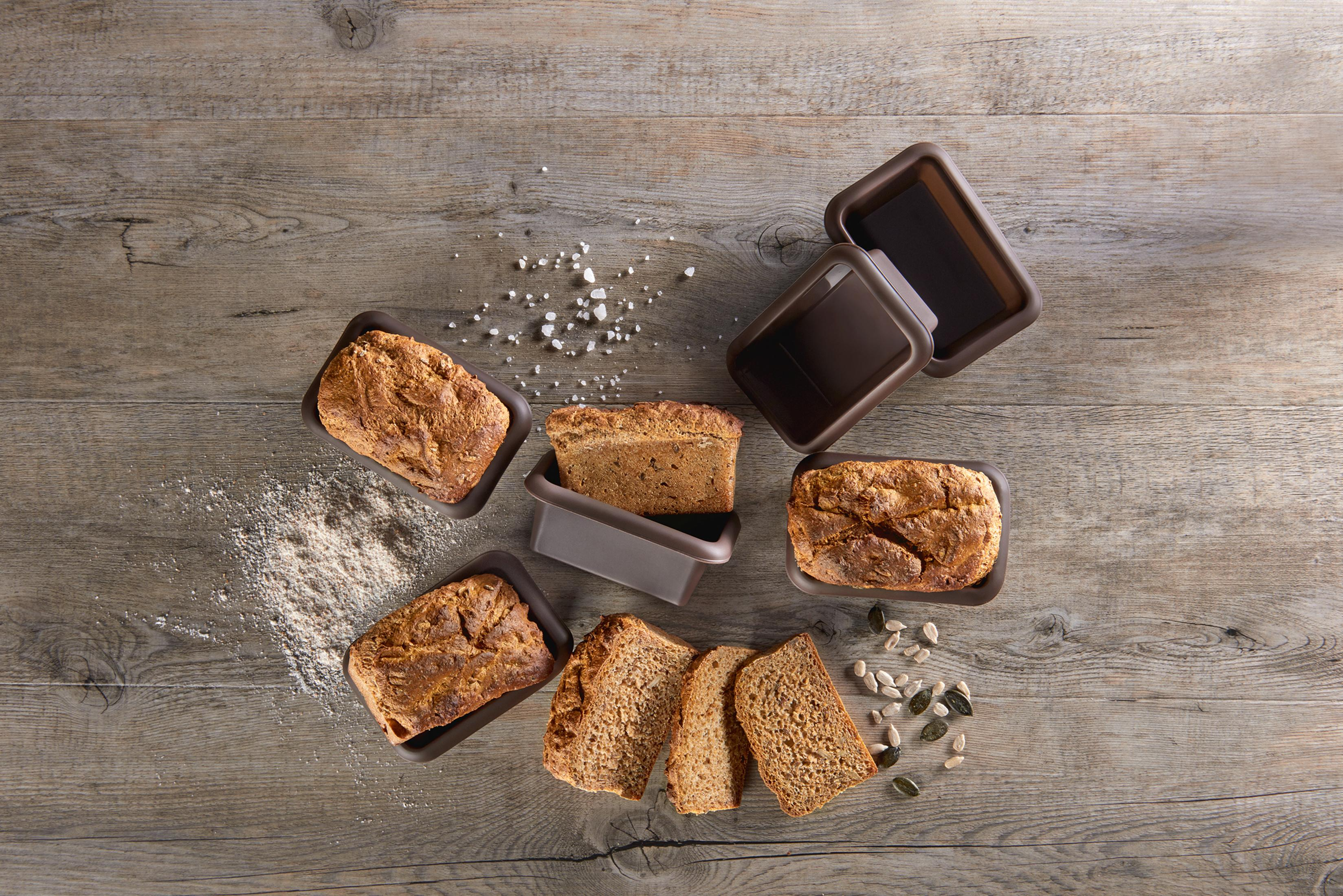Drewniany stół z bochenkami chleba, pojemnikiem, mąką i nasionami.