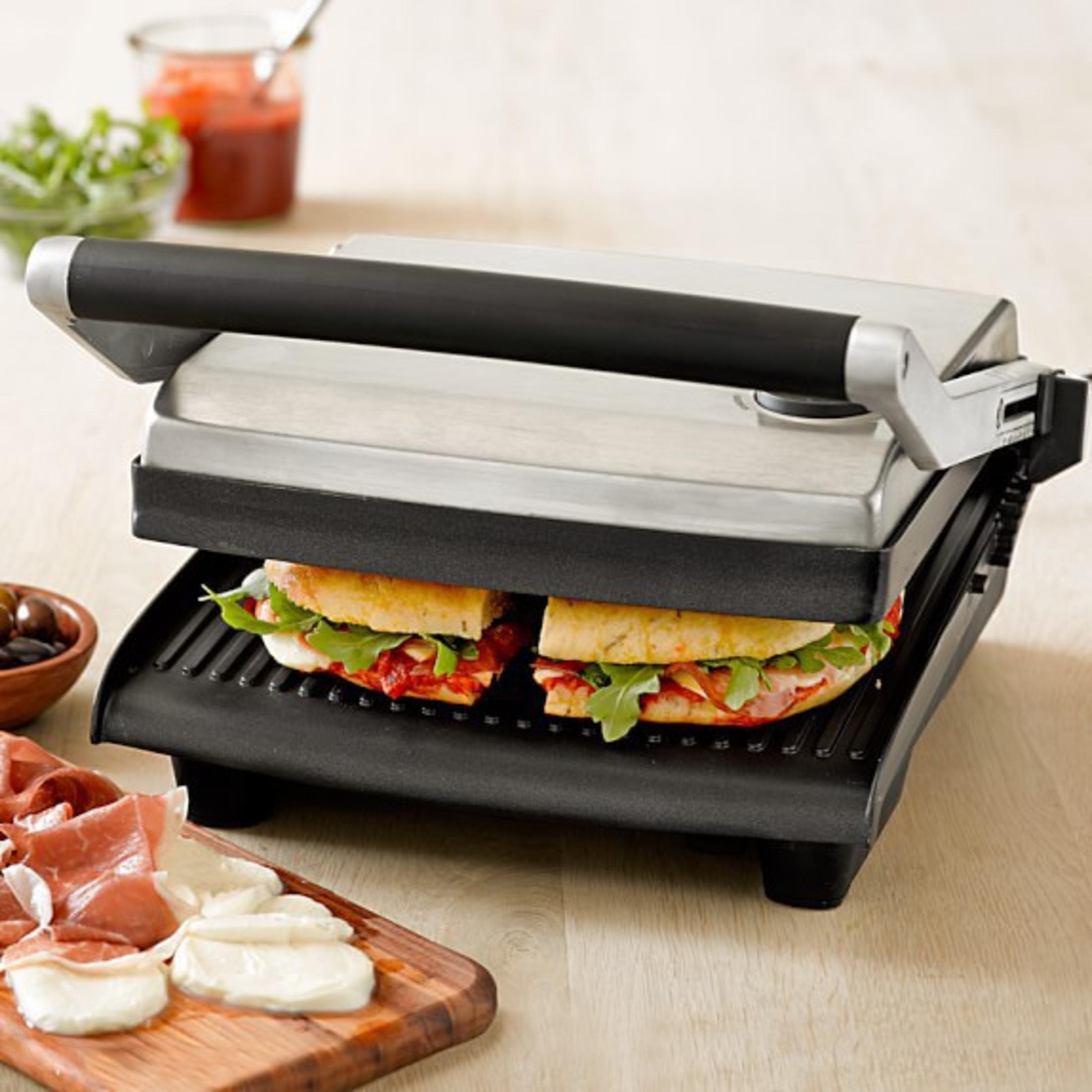 Eine Panini-Presse mit zwei Sandwiches darin, zusammen mit Zutaten wie Oliven, Schinken und Grünzeug.